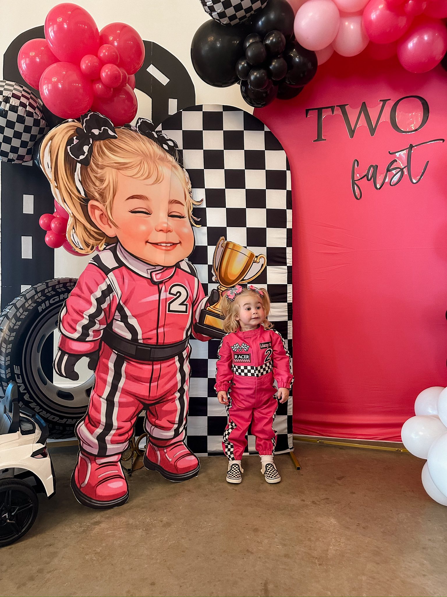 Toddler wearing a pink race car driver jumpsuit at a Two Fast themed birthday party with a matching cutout display.