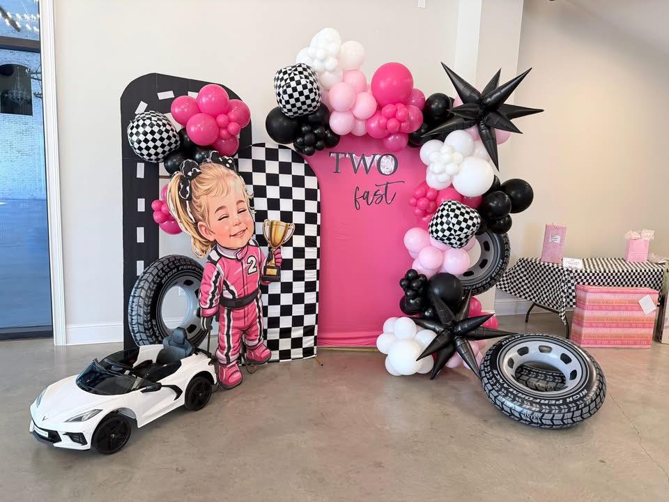 Two Fast themed birthday party backdrop with pink and black balloons, checkered patterns, and a race car cutout.