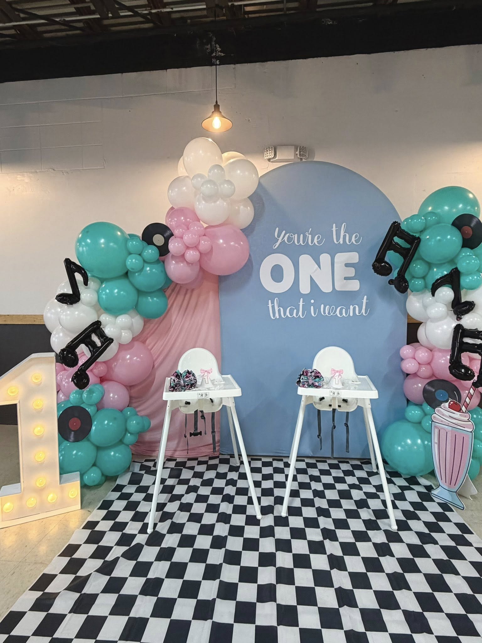 1950s rock and roll themed first birthday party decoration with a balloon arch, light-up number one, and high chairs