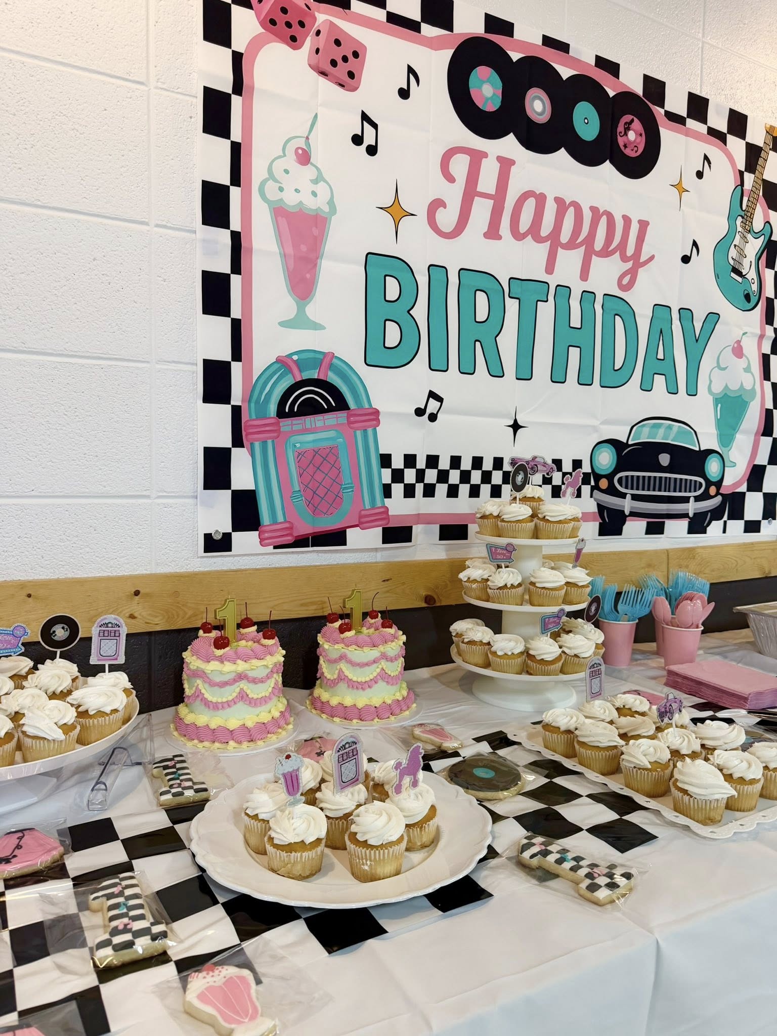 1950s rock and roll themed birthday party setup with cupcakes, two small cakes, and a Happy Birthday wall banner