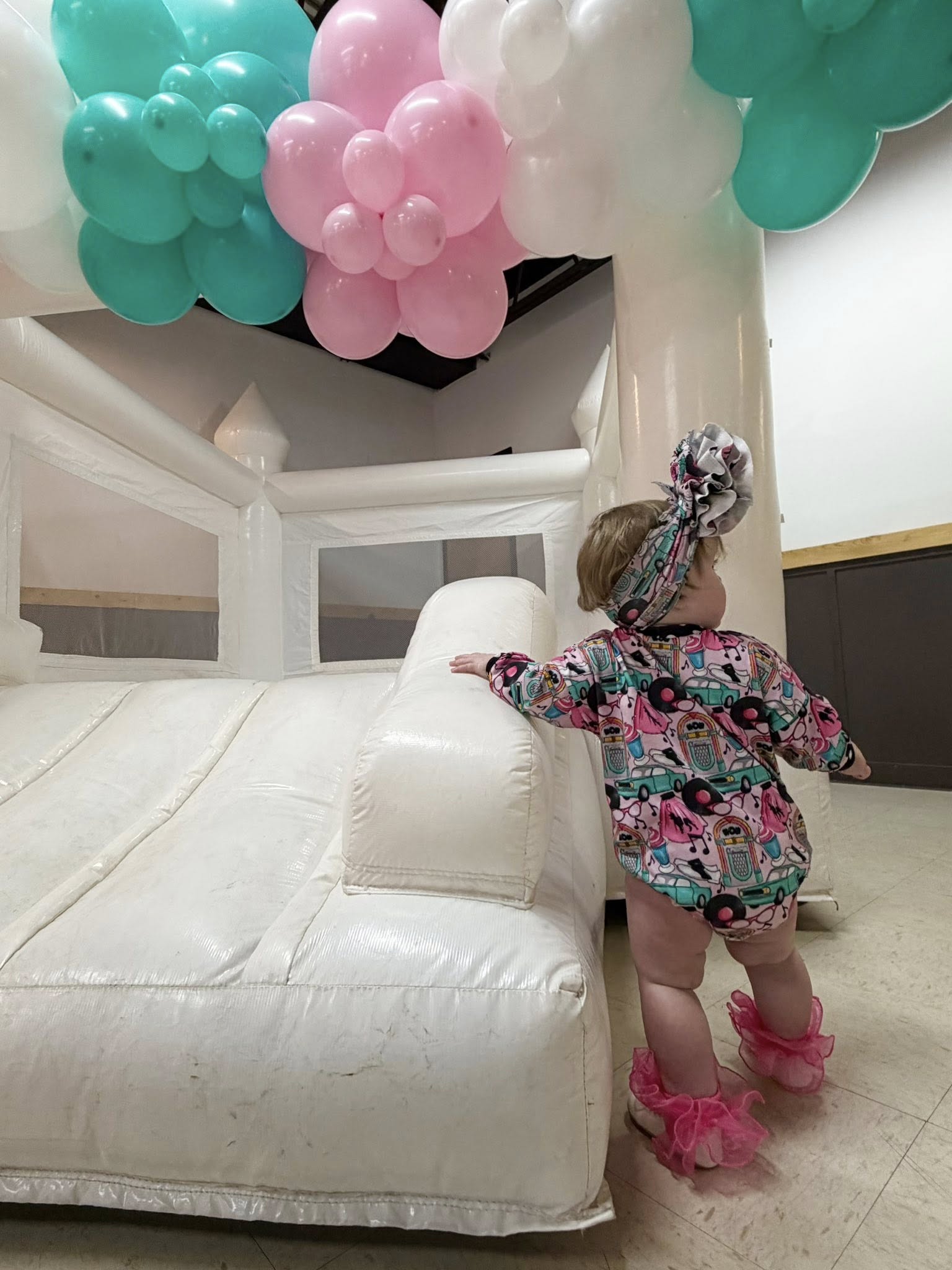 Toddler wearing a patterned romper and ruffled headband standing next to a white indoor bounce house.
