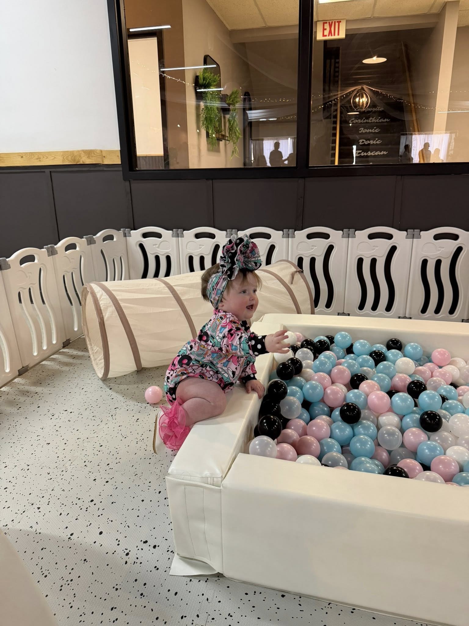 A smiling baby with a large bow playing in a soft white ball pit filled with pink, blue, and black balls.