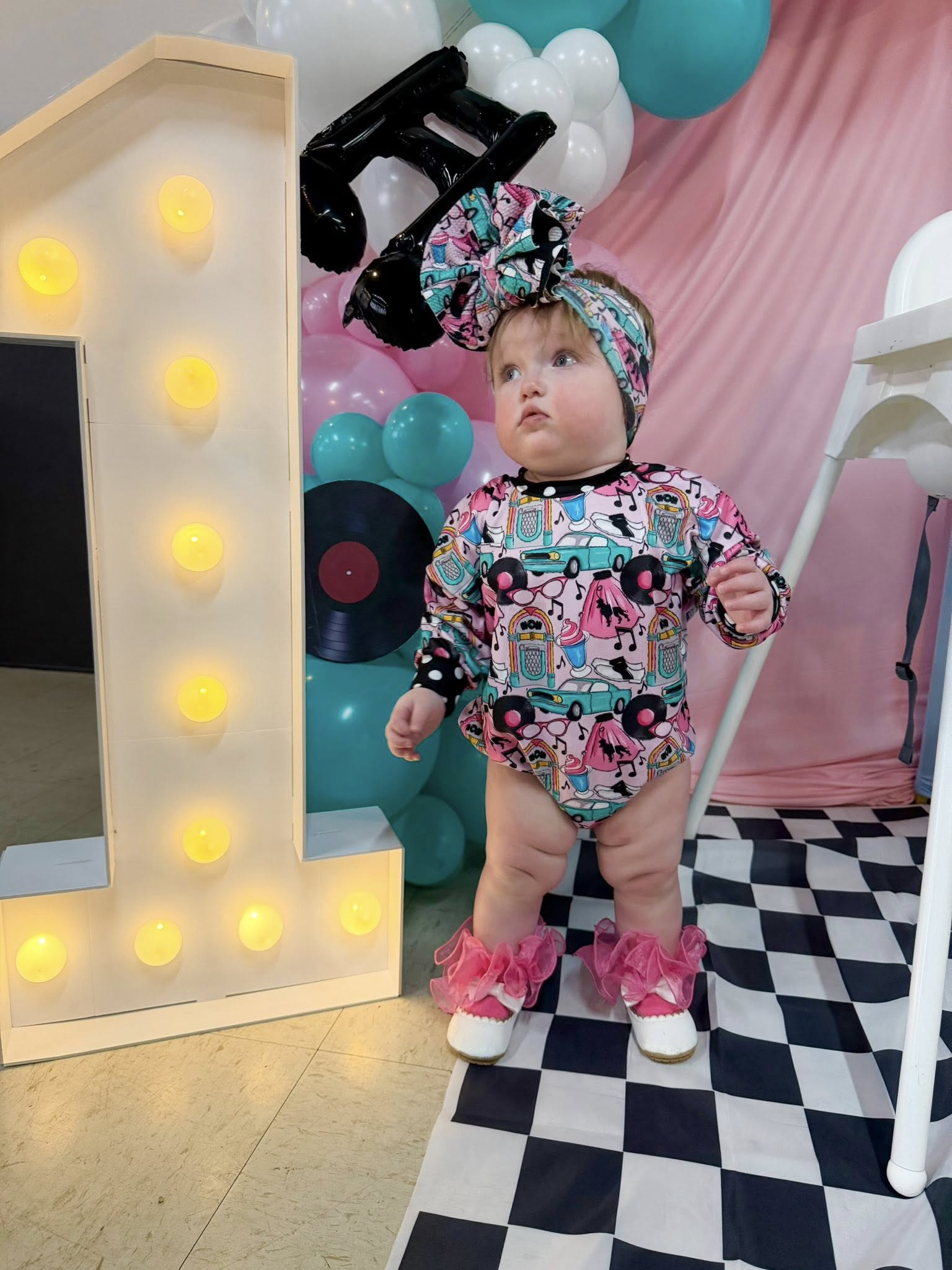 Toddler wearing a 1950s rock and roll themed bodysuit and headband standing next to a large light-up number one.