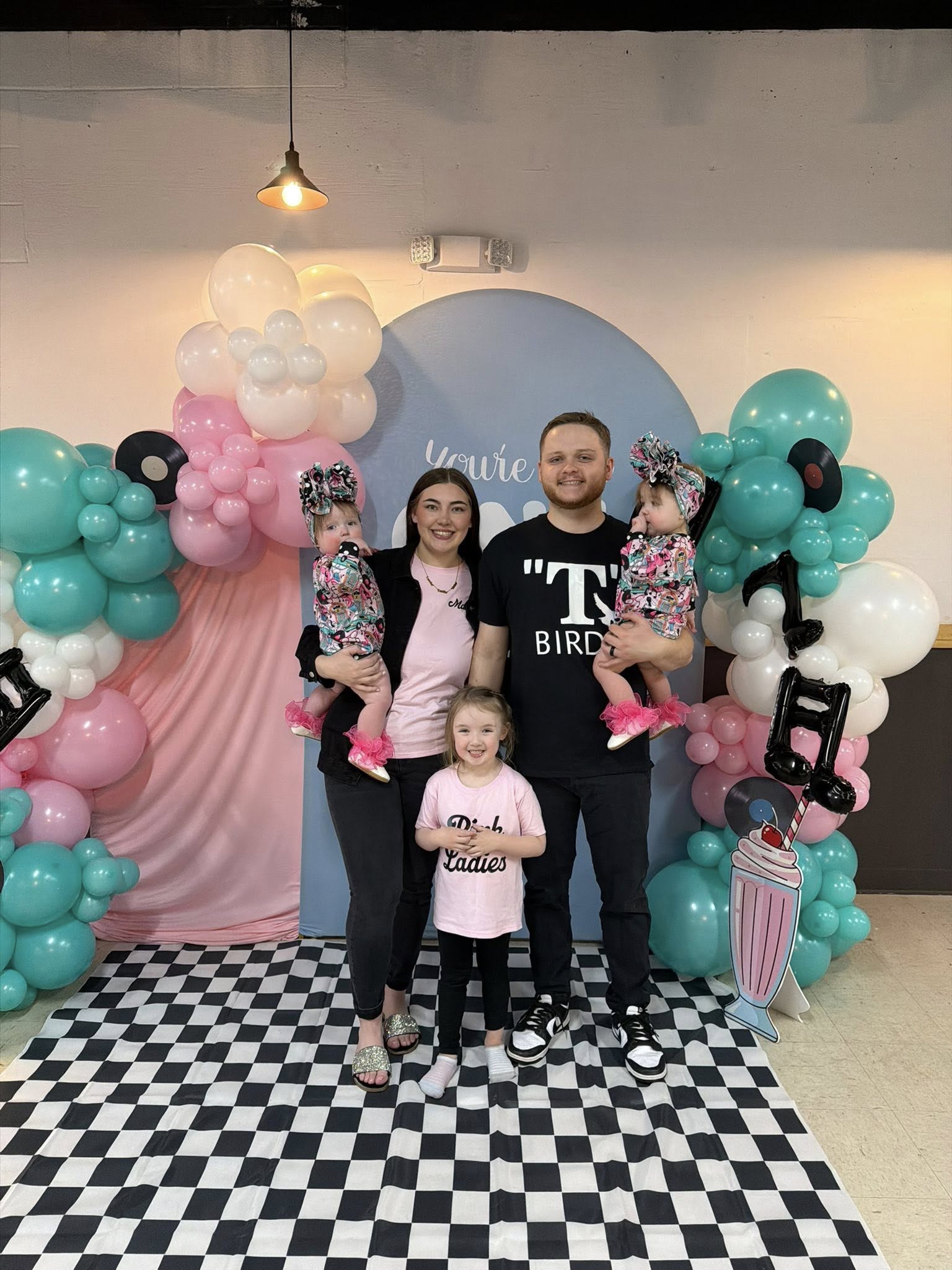 Family posing in 1950s Grease-themed outfits for a photoshoot with balloons and a checkered party backdrop