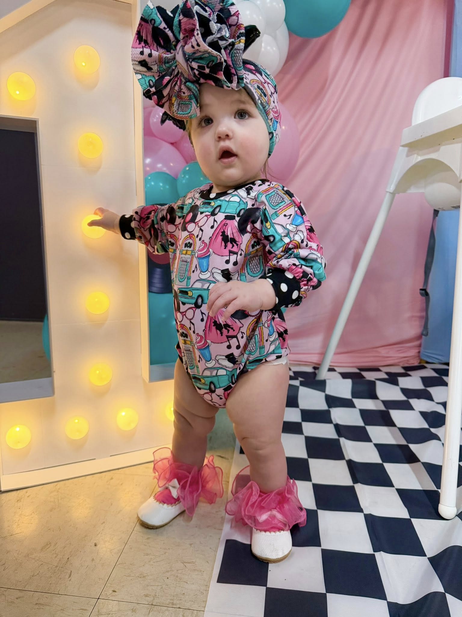 Toddler wearing 1950s themed long-sleeve romper with matching bow and frilly pink anklets for a first birthday party.