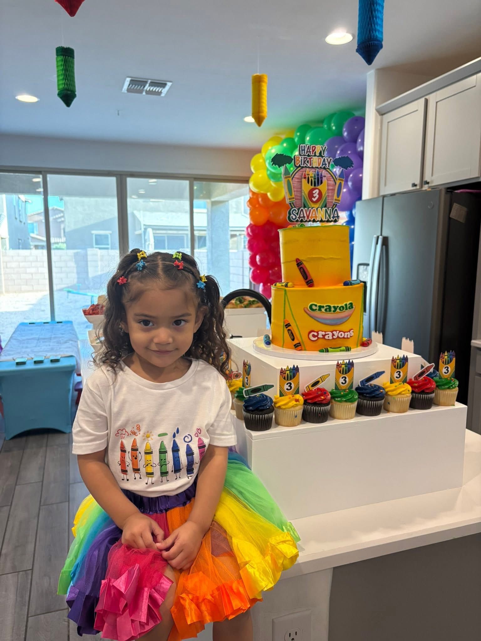 Young girl in a rainbow outfit at a Crayola-themed birthday party next to a two-tier yellow birthday cake.