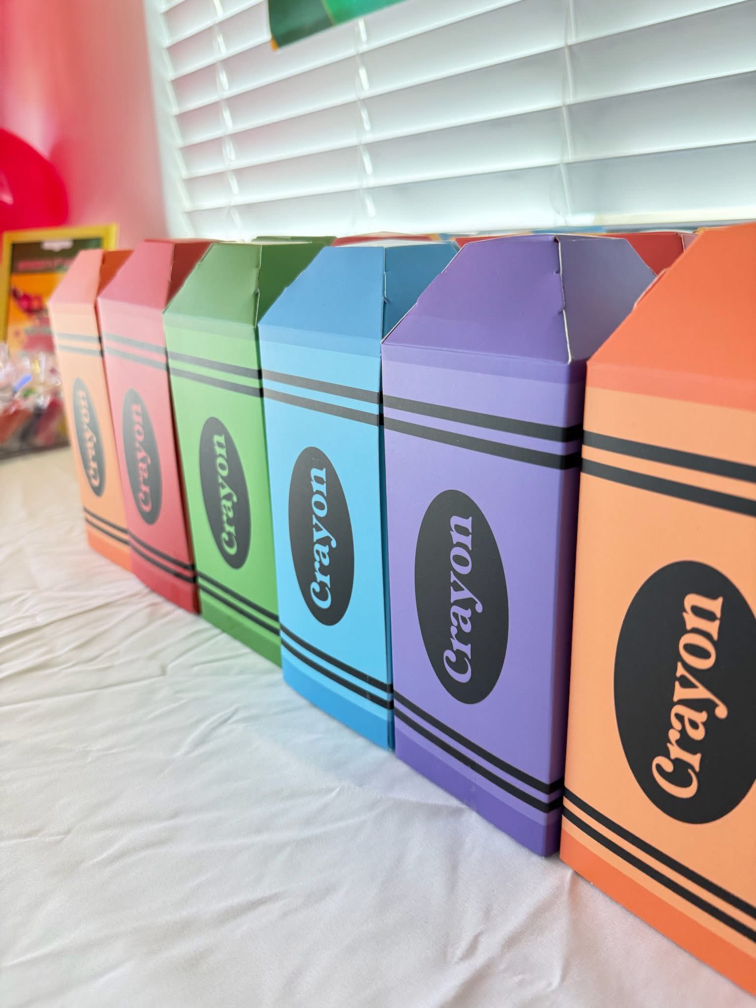 Row of colorful cardboard crayon-shaped favor boxes on a white tablecloth