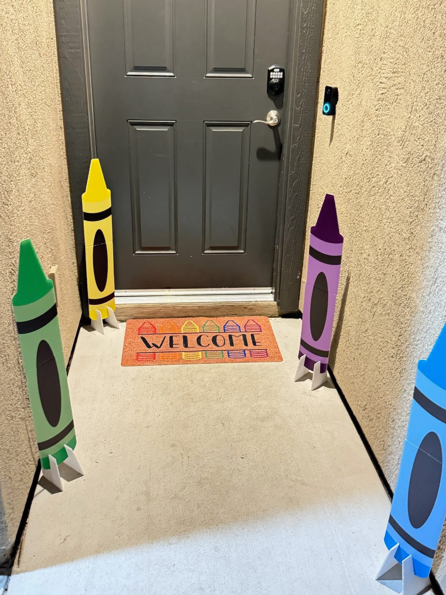 Front porch decor featuring four colorful giant crayon standups and a Welcome mat with crayon illustrations.