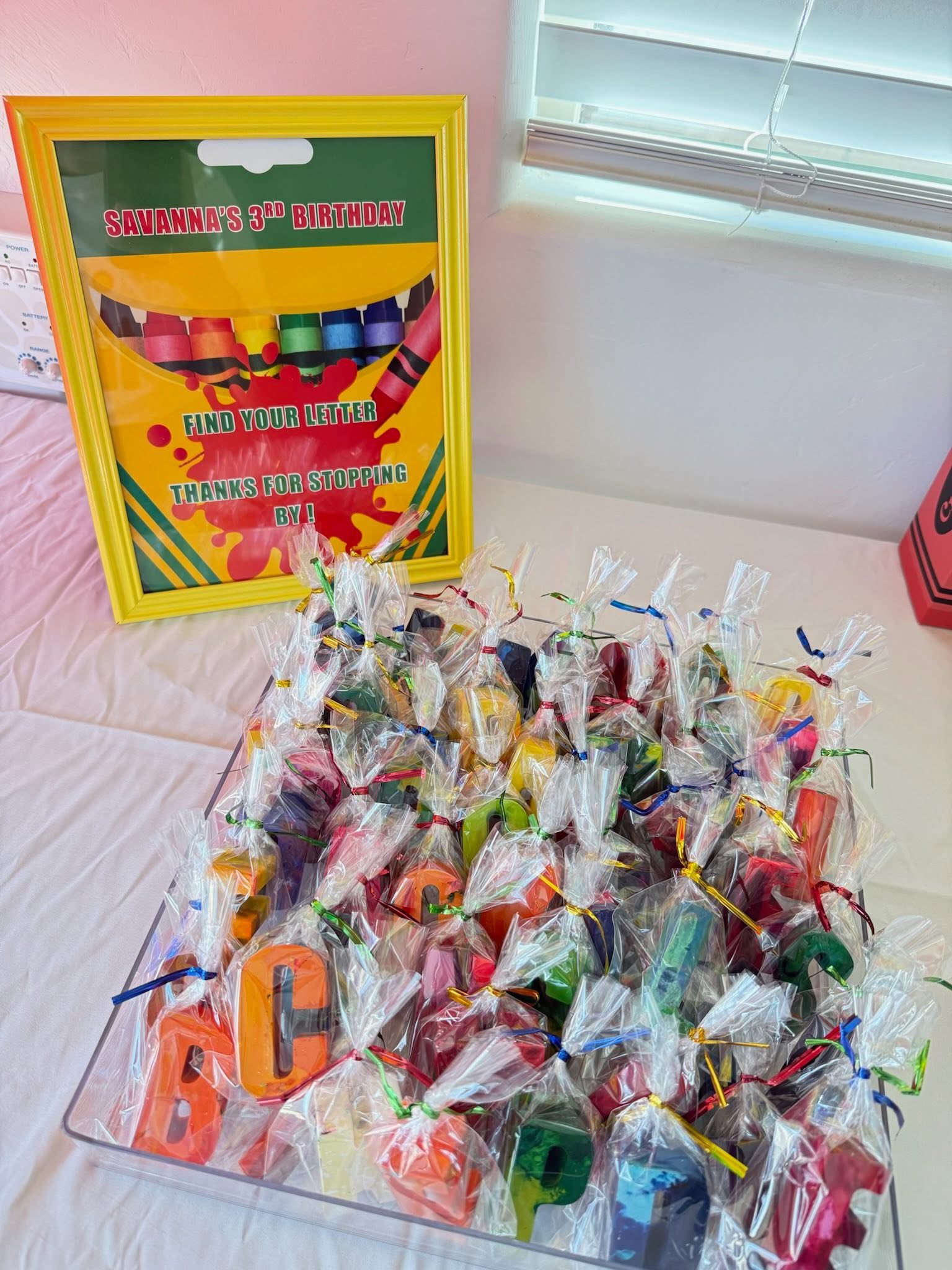 Table display for a 3rd birthday party featuring individual cellophane-wrapped letter crayons in a clear bin.