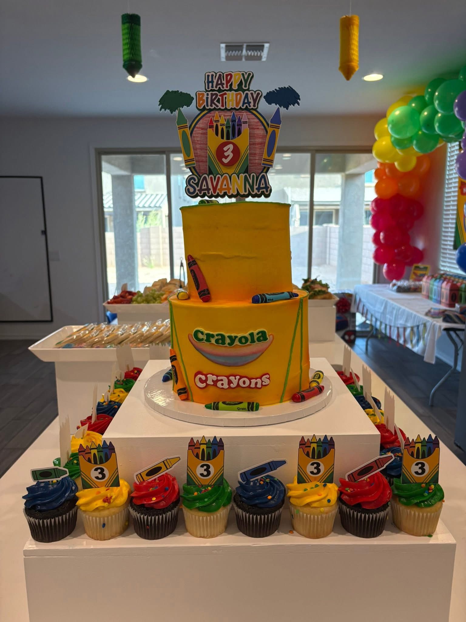 Two-tier yellow Crayola themed birthday cake with a number 3 topper for Savanna, surrounded by colorful cupcakes.