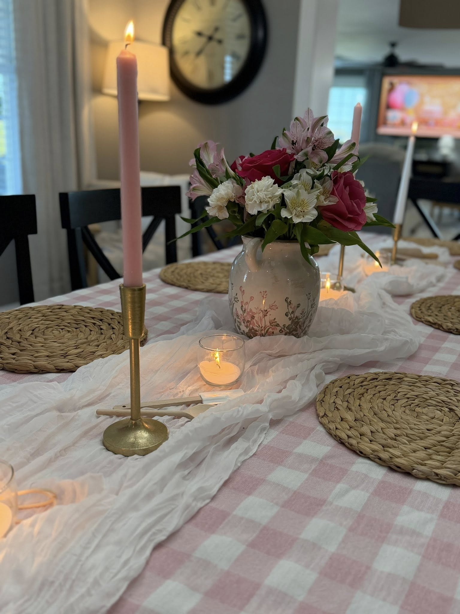 Dining table set with pink gingham tablecloth, woven placemats, and floral centerpiece with candles