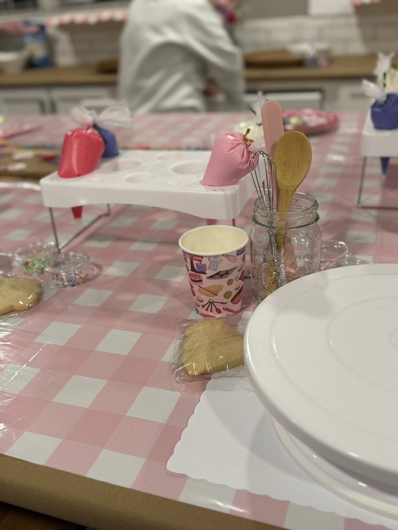 Decorated cookie party setup with piping bags, utensils in a mason jar, cookie cutters, and paper cups on a pink gingham tablecloth
