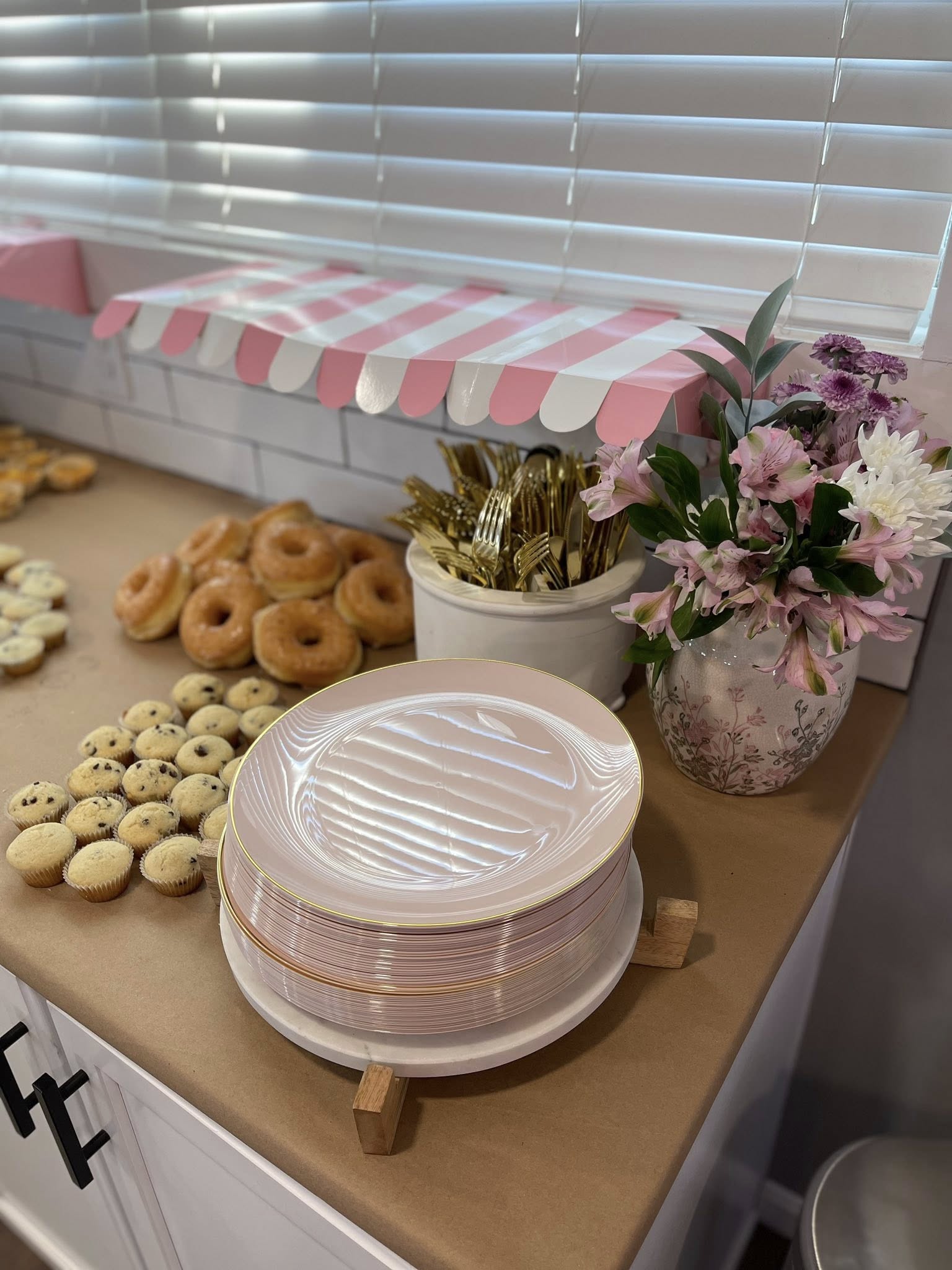 Table setting with stack of blush pink plates, assorted donuts, mini muffins, gold cutlery, and floral arrangement.
