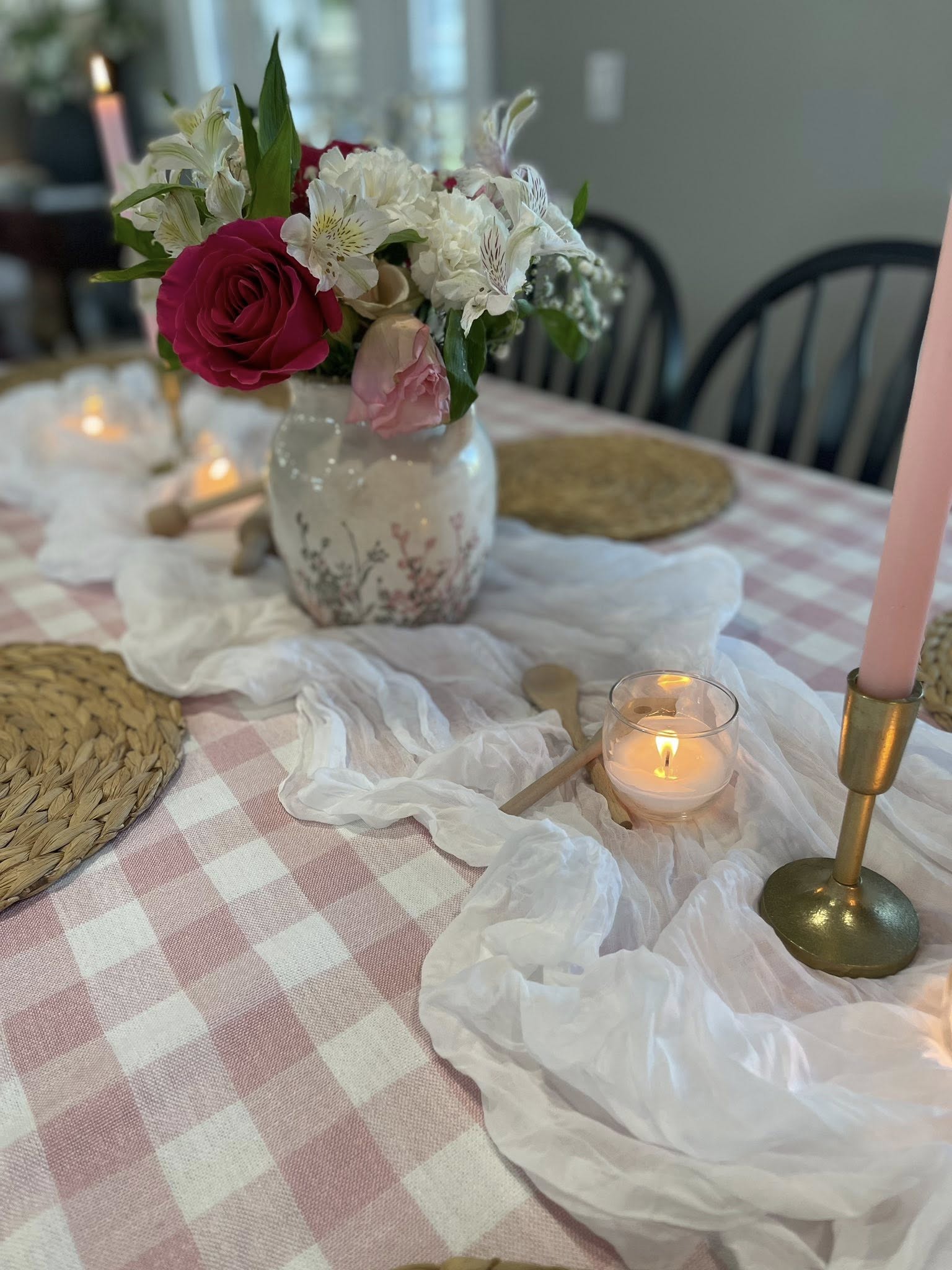 Dining table set for a special occasion with a pink checkered runner, floral vase, candles, and woven placemats.