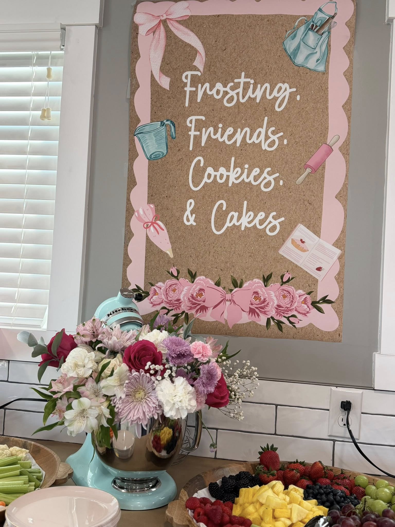Decorative sign on a wall reading Frosting, Friends, Cookies, & Cakes above a floral arrangement and fruit platter.