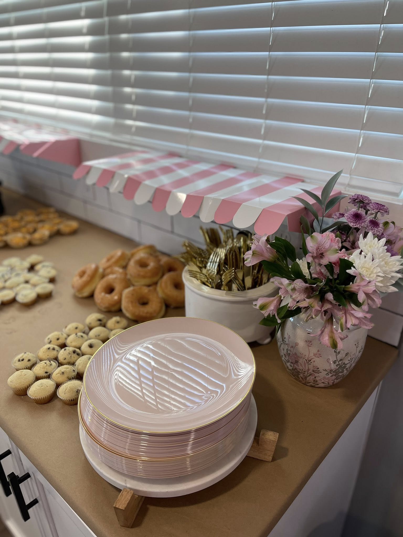 Table spread featuring a stack of blush pink plates with gold rims, glazed donuts, mini muffins, and a floral bouquet.