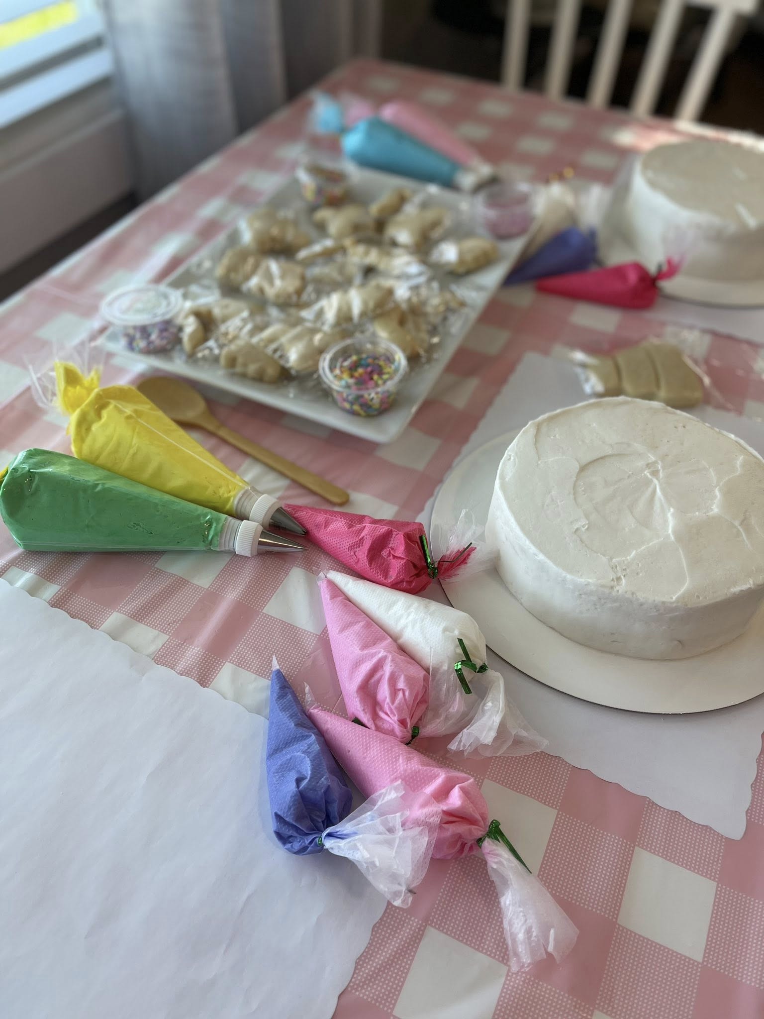 Table set up for cake and cookie decorating with various colored icing bags, plain cakes, and sprinkles.