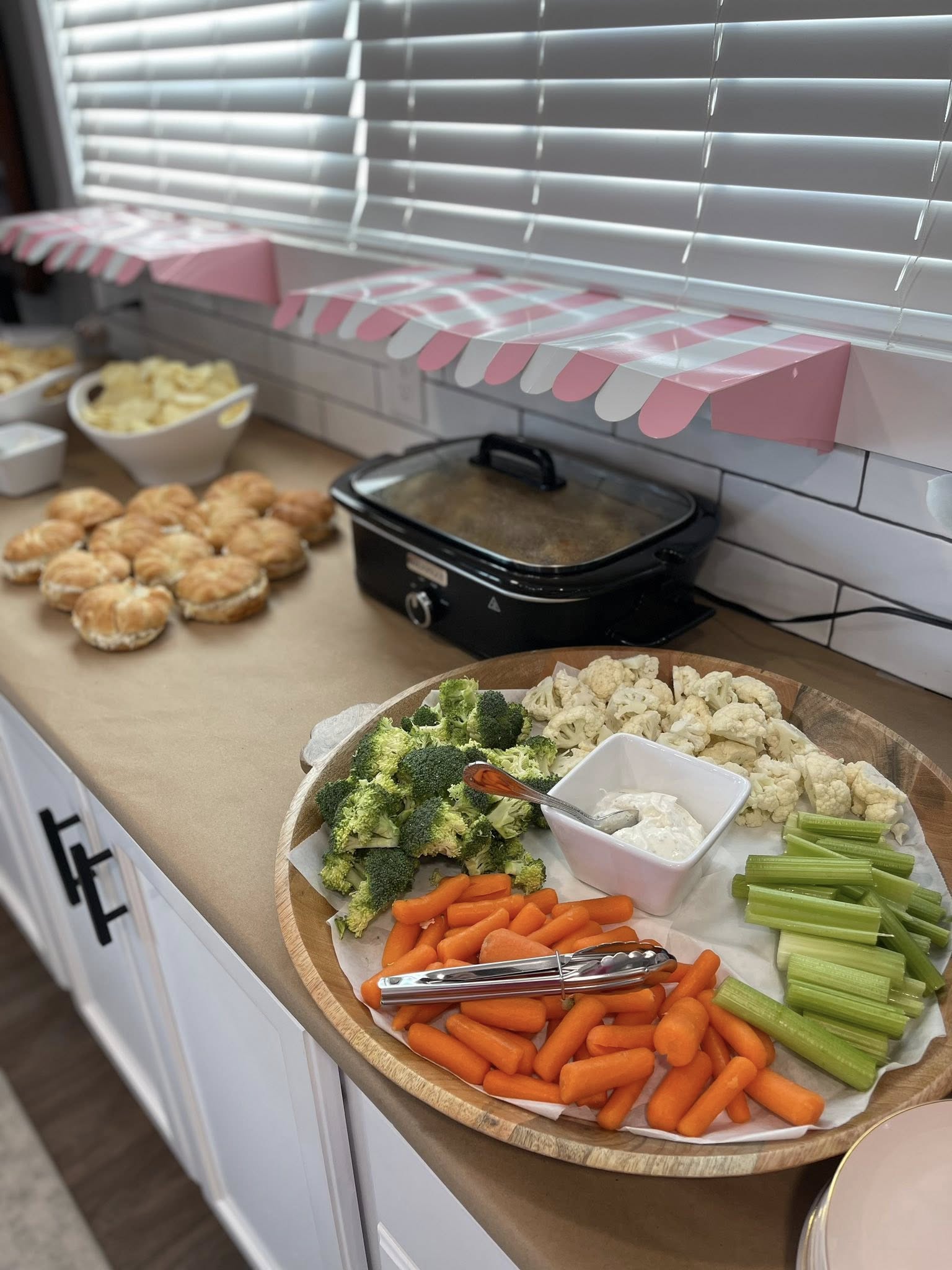 A party grazing table featuring a wooden tray of fresh vegetables with dip, buns, and a slow cooker.