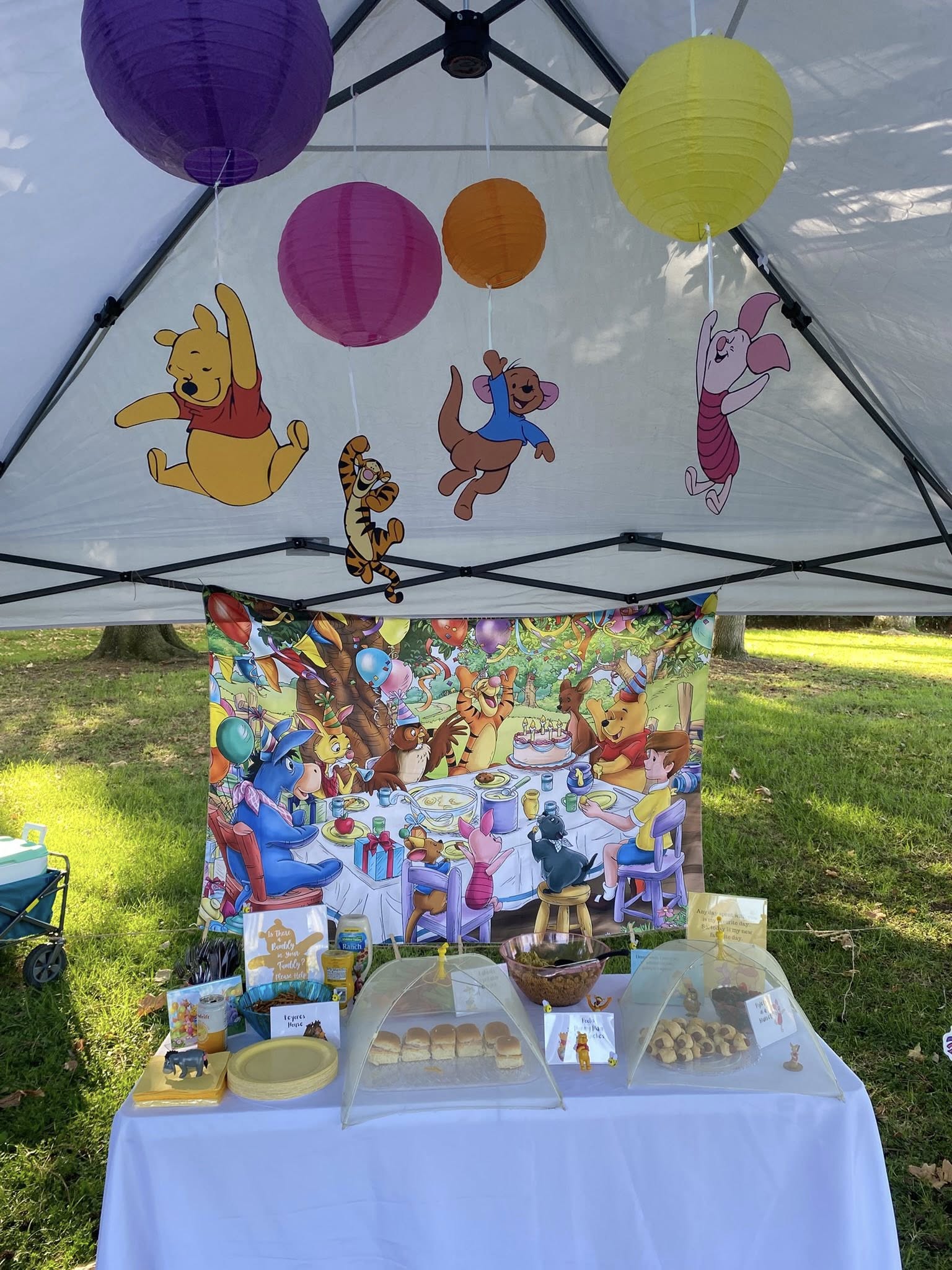 Winnie the Pooh themed party table outdoors with a decorated canopy and character-themed snacks.