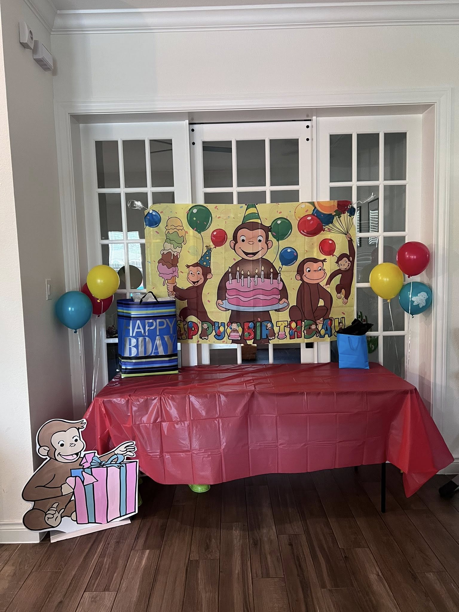 Curious George happy birthday party banner with matching balloons and gift bags on a red table.