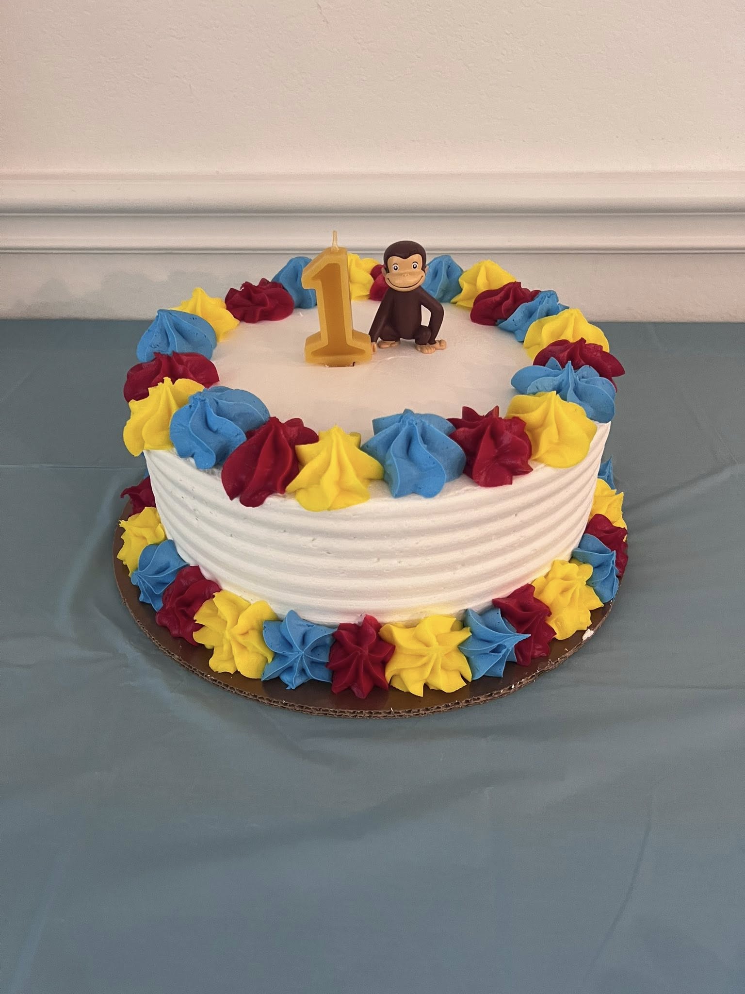 First birthday cake decorated with blue, yellow, and red rosettes and a Curious George topper with a number one candle.