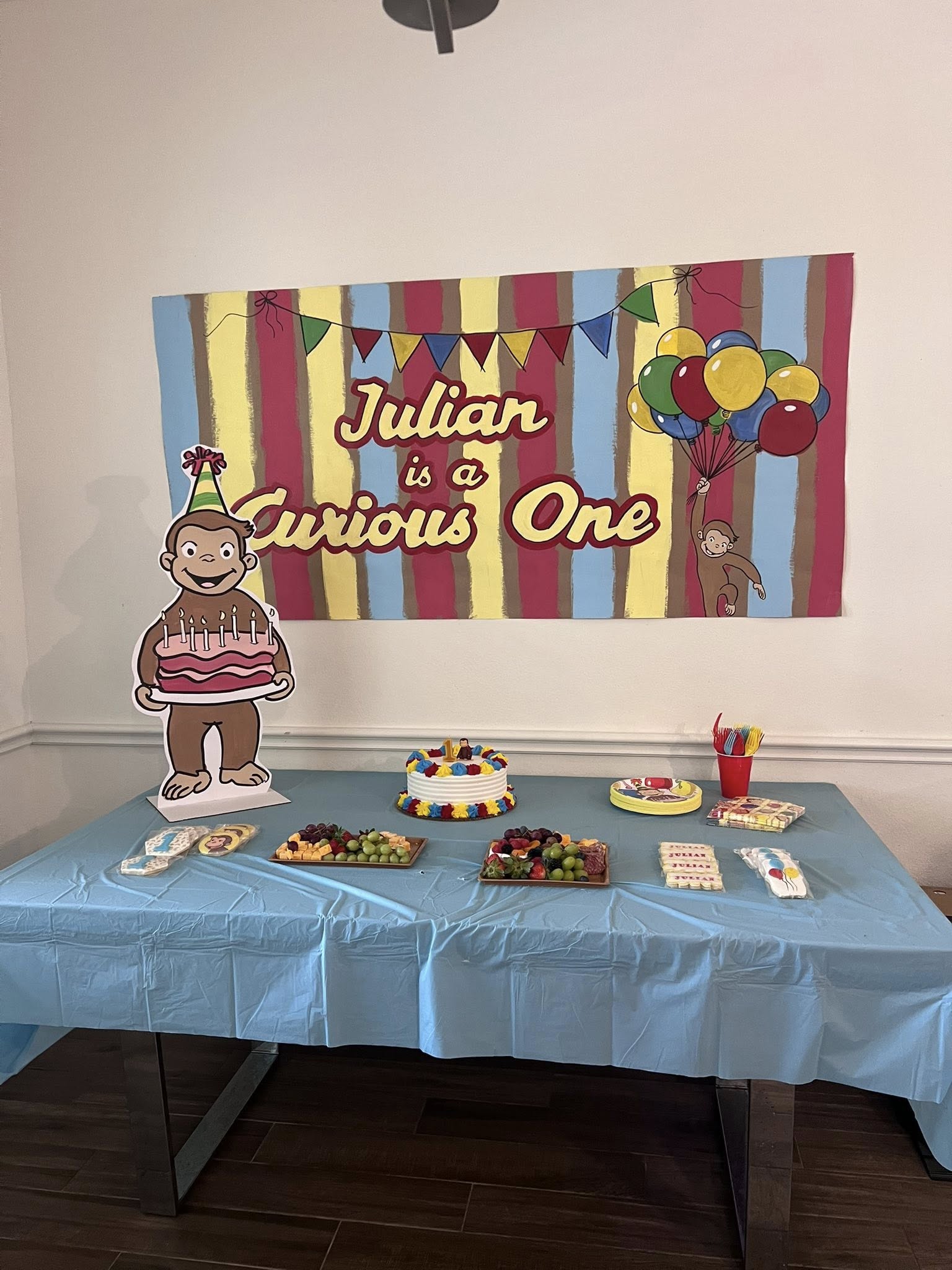 Curious George themed first birthday party table with cake, fruit platters, and custom banner for Julian.