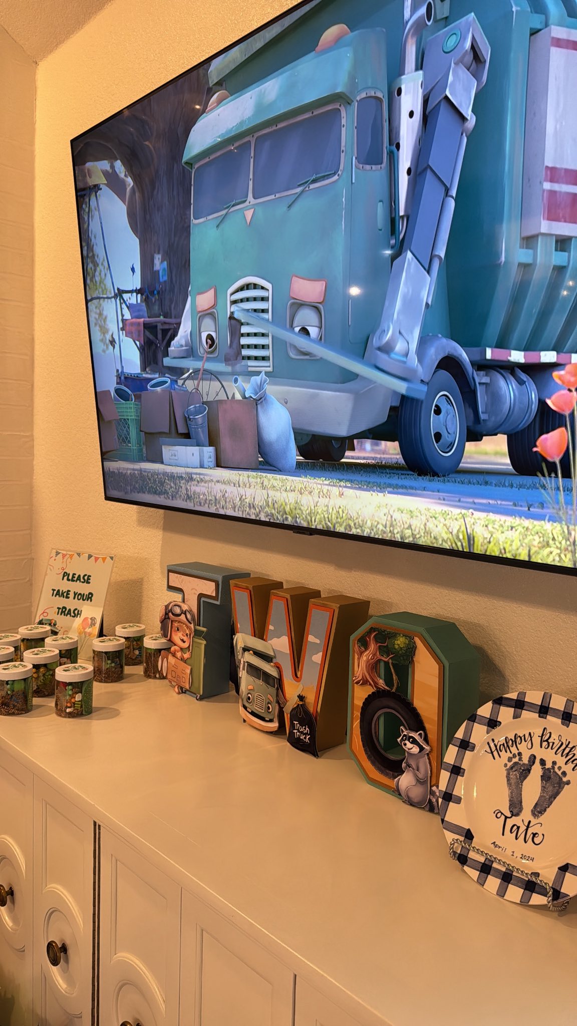 Trash Truck themed second birthday party decorations with custom lettering, a birthday plate, and jars on a console table under a TV screen showing the show.