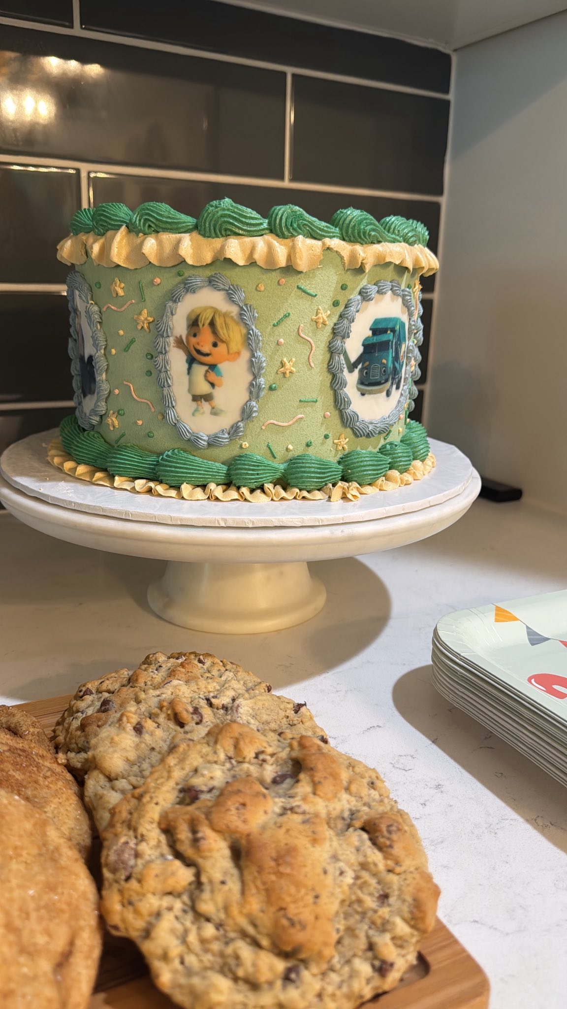 Custom birthday cake with green frosting and edible character images on a cake stand next to chocolate chip cookies