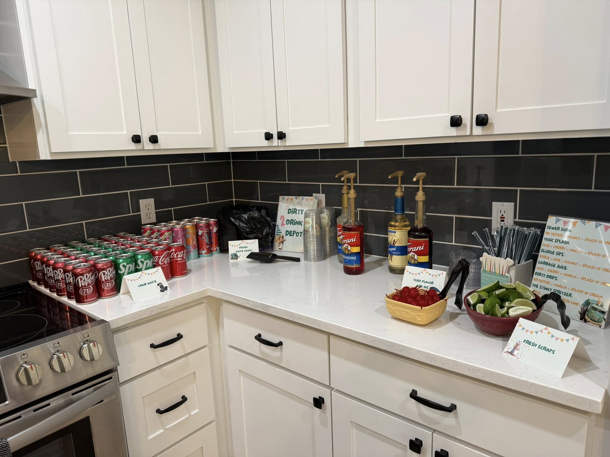 DIY soda bar setup on a kitchen counter featuring various sodas, Torani syrups, cherries, limes, and custom drink signs.