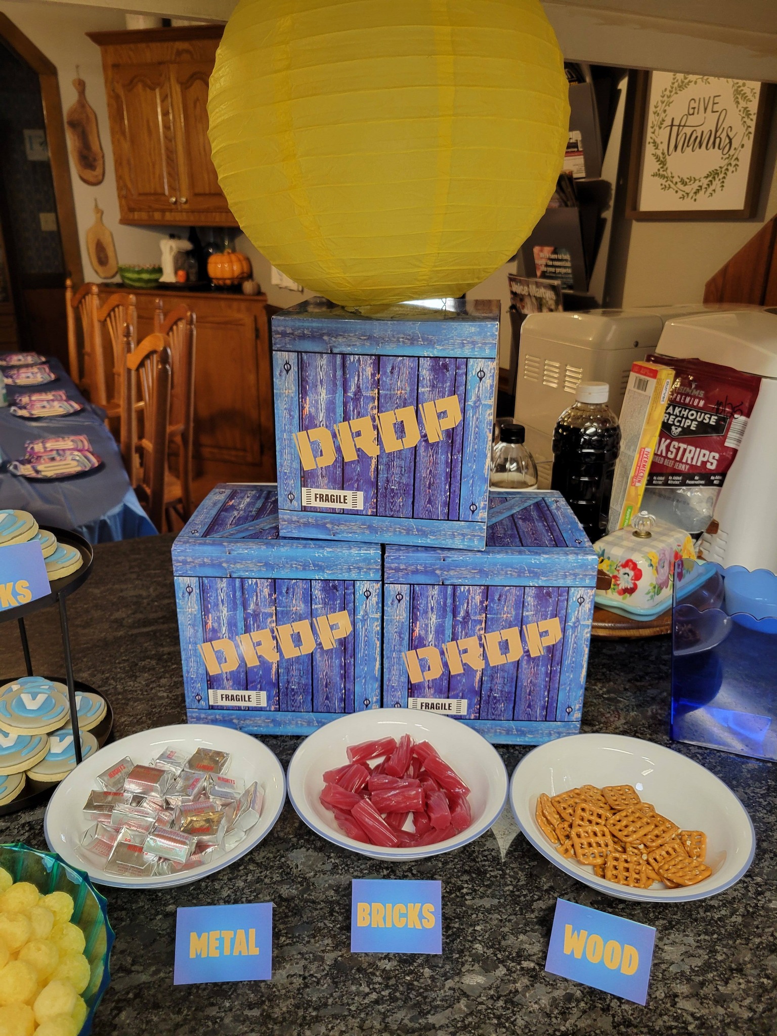Party game setup with three blue crates labeled Drop and bowls of candy, bricks and wood labeled snacks.