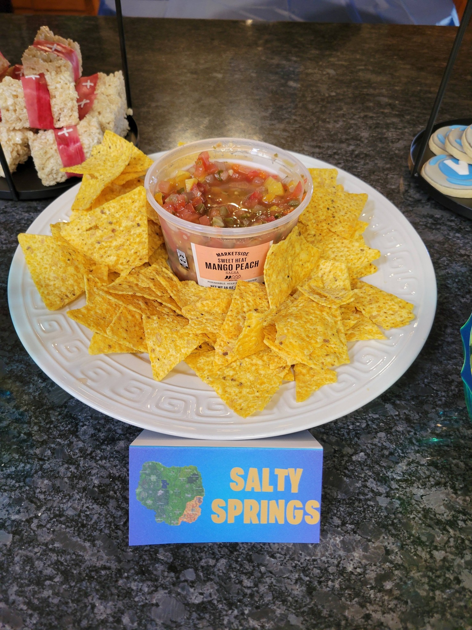 A white plate heaped with tortilla chips surrounding a container of Marketside Sweet Heat Mango Peach salsa, labeled Salty Springs.