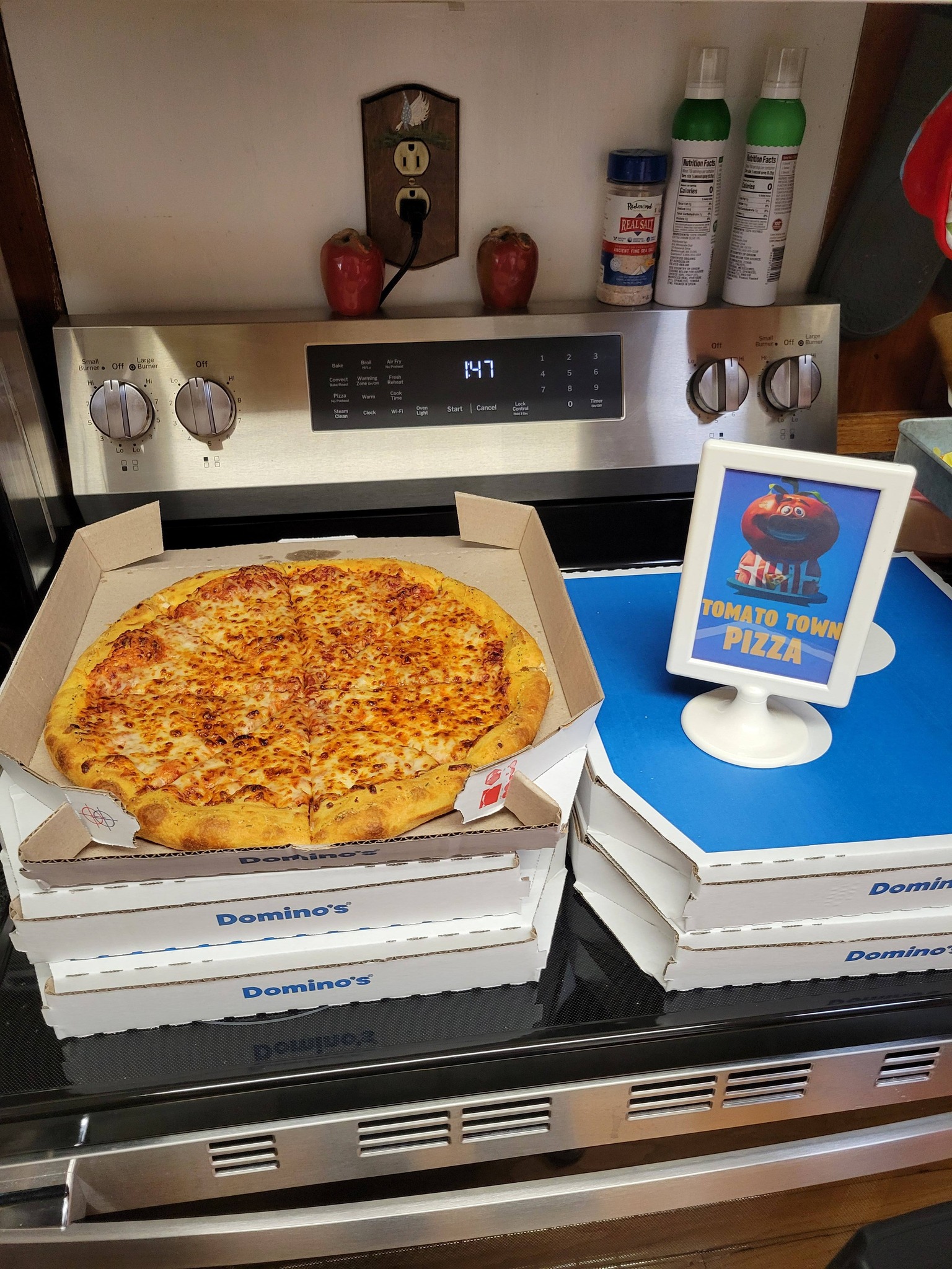 A cheese pizza in a Domino's box next to a Tomato Town Pizza sign, sitting on a kitchen stove.