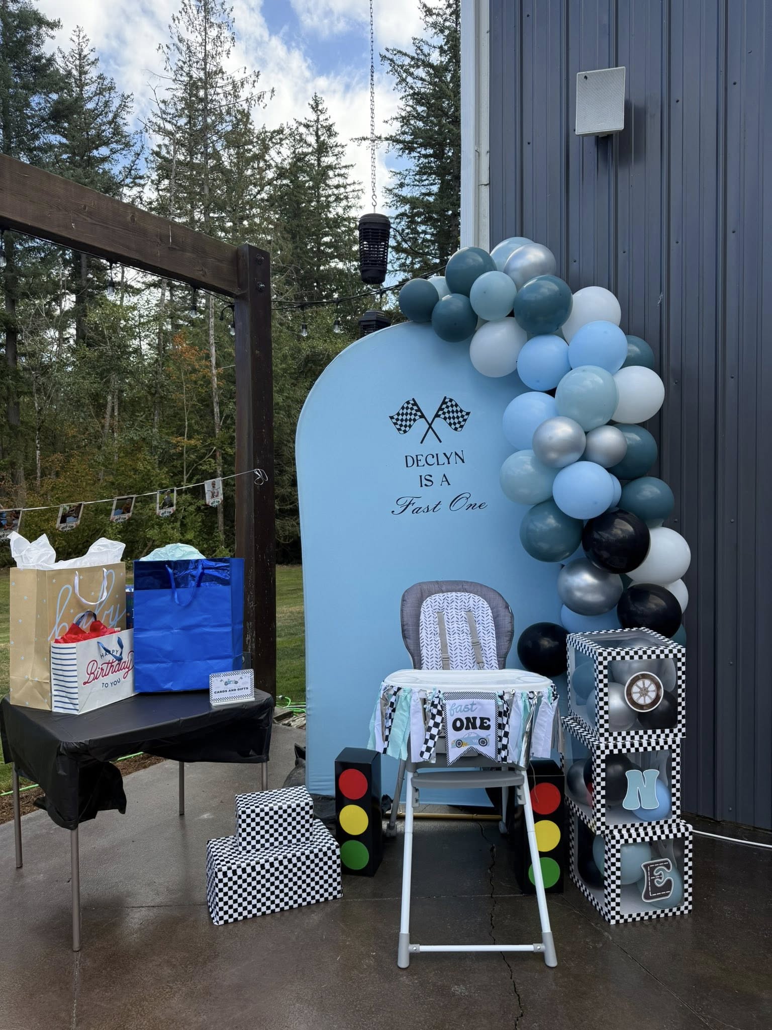 Race car-themed first birthday party decorations, including a blue backdrop, balloon arch, high chair, and gift table.