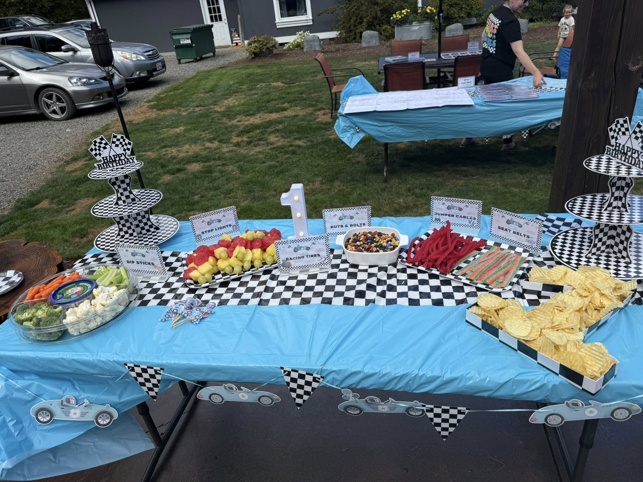Race car-themed first birthday party snack table with checkered decorations and creative food labels.