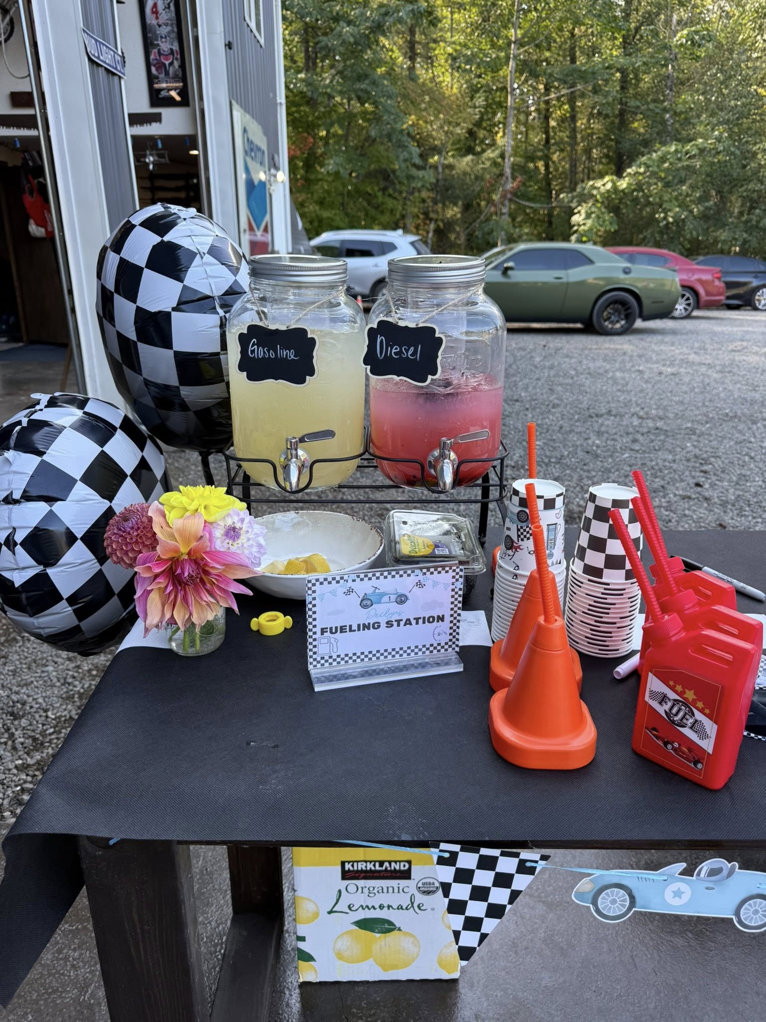 Race car-themed drink station with beverage dispensers labeled Gasoline and Diesel, checkered balloons, and race cones.