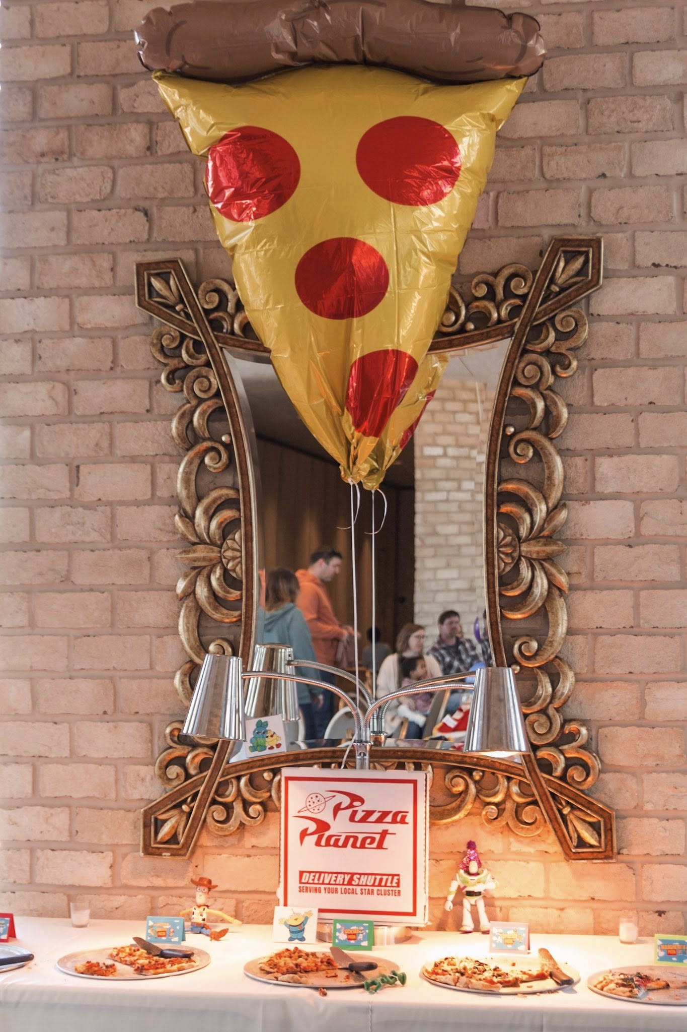 Pizza Planet themed buffet table featuring a pizza slice balloon, character figurines, and serving plates.