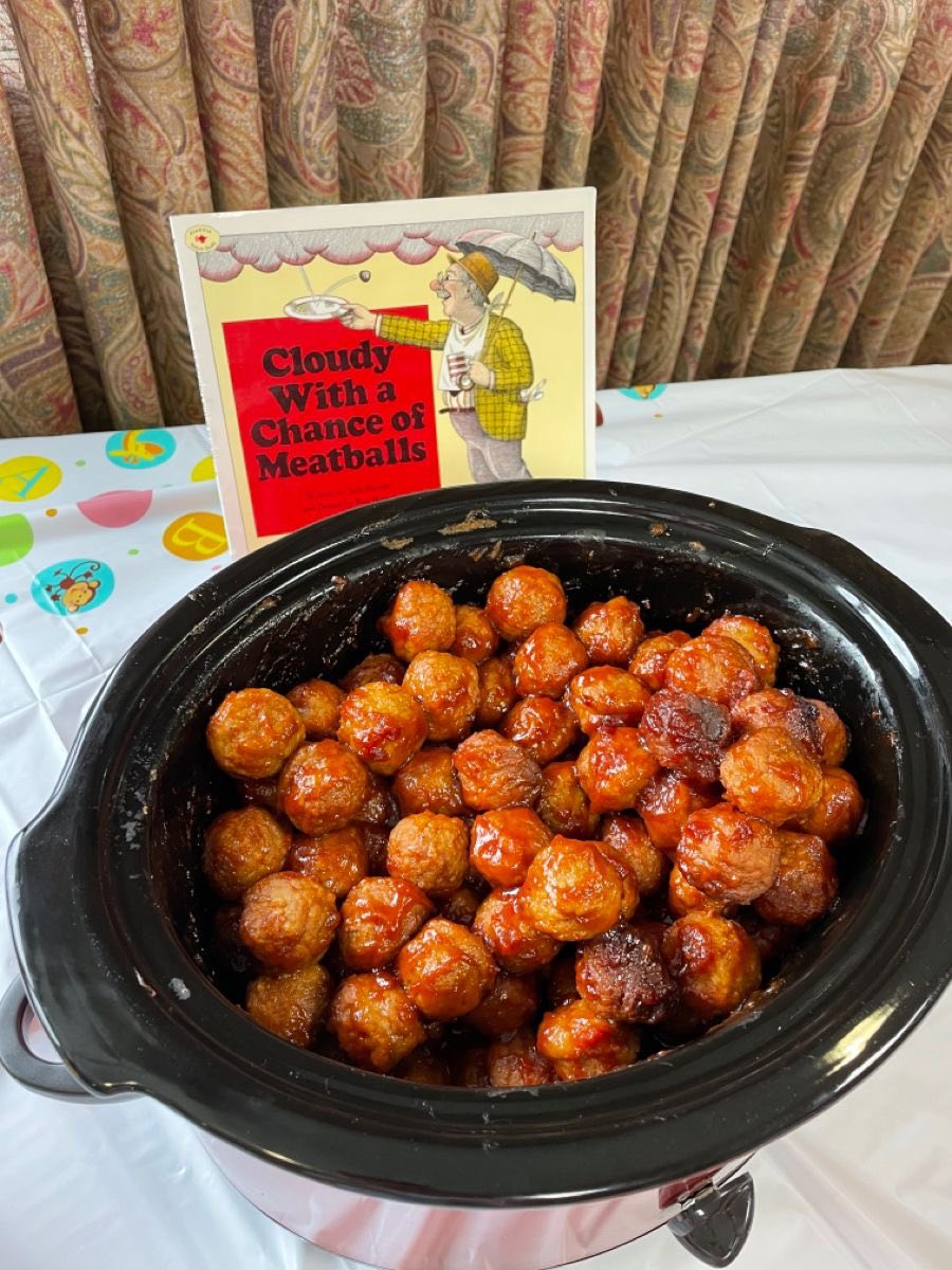 Slow cooker filled with glazed meatballs displayed next to the book Cloudy With a Chance of Meatballs