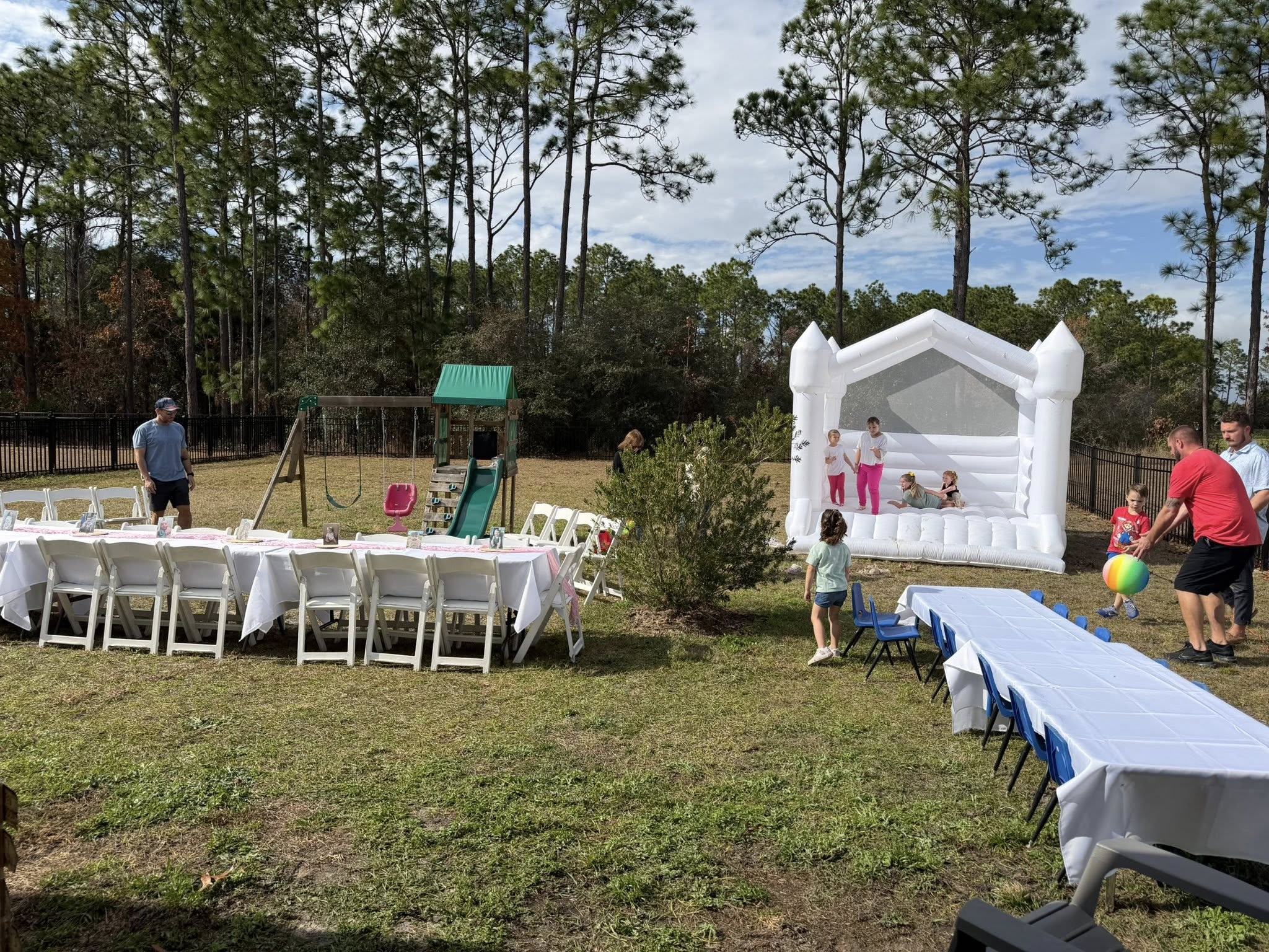 Outdoor birthday party setup with a white bounce house, dining tables, and a swing set in a backyard.