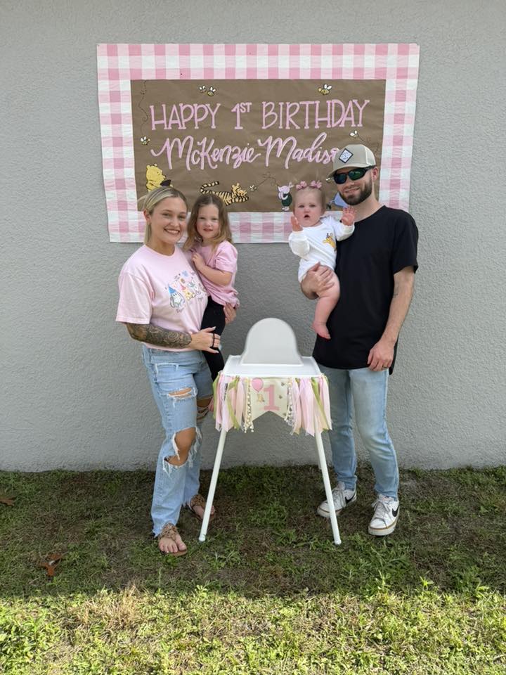 Family standing in front of a Winnie the Pooh themed first birthday backdrop with a high chair decorated with ribbons.