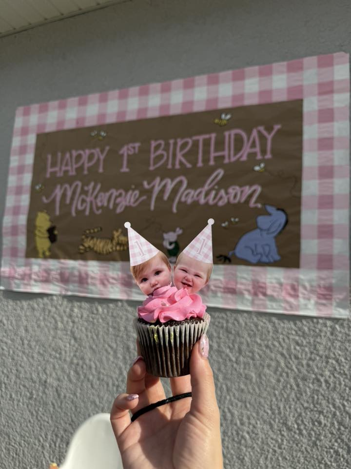 Cupcake topped with custom face cake toppers in party hats, held in front of a First Birthday banner.