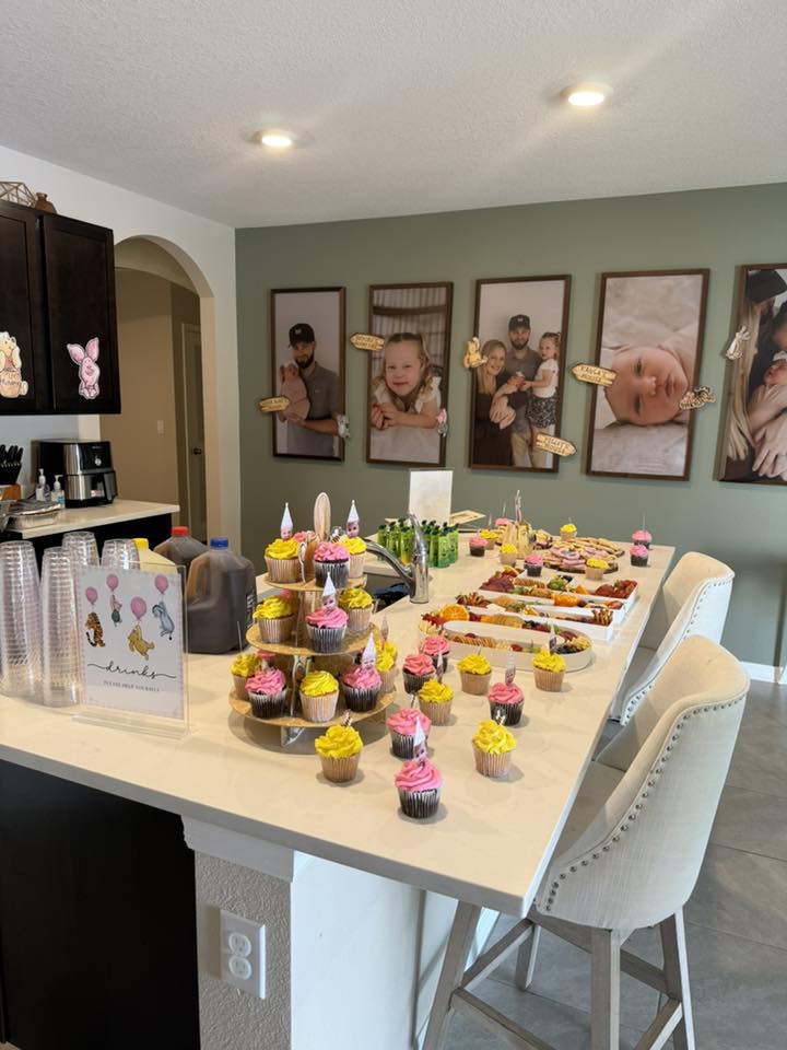 Kitchen counter set up for a party with a cupcake tower and assorted snacks under family portraits on the wall.