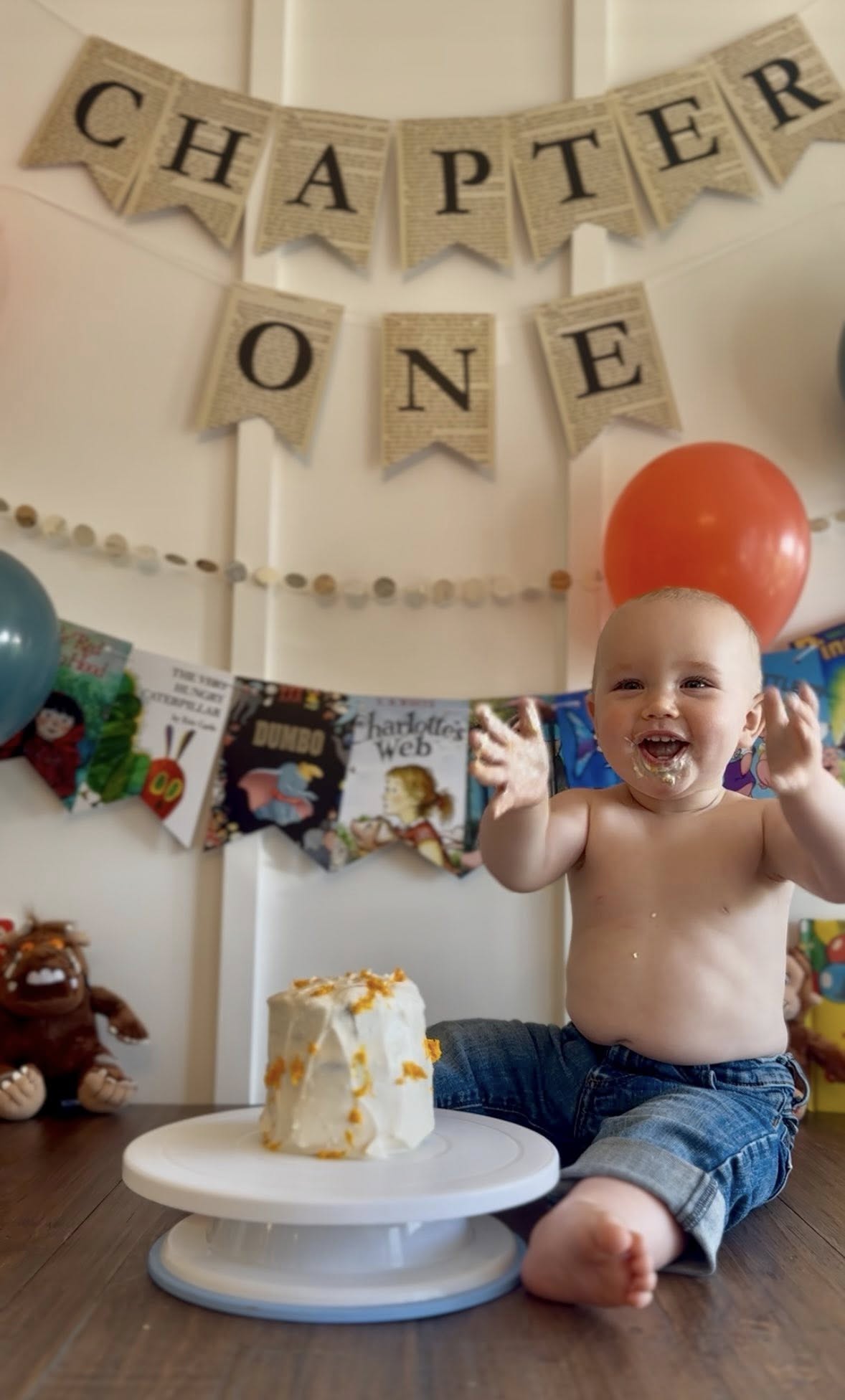 Happy baby sitting in front of a smash cake with a book-themed first birthday party decoration background