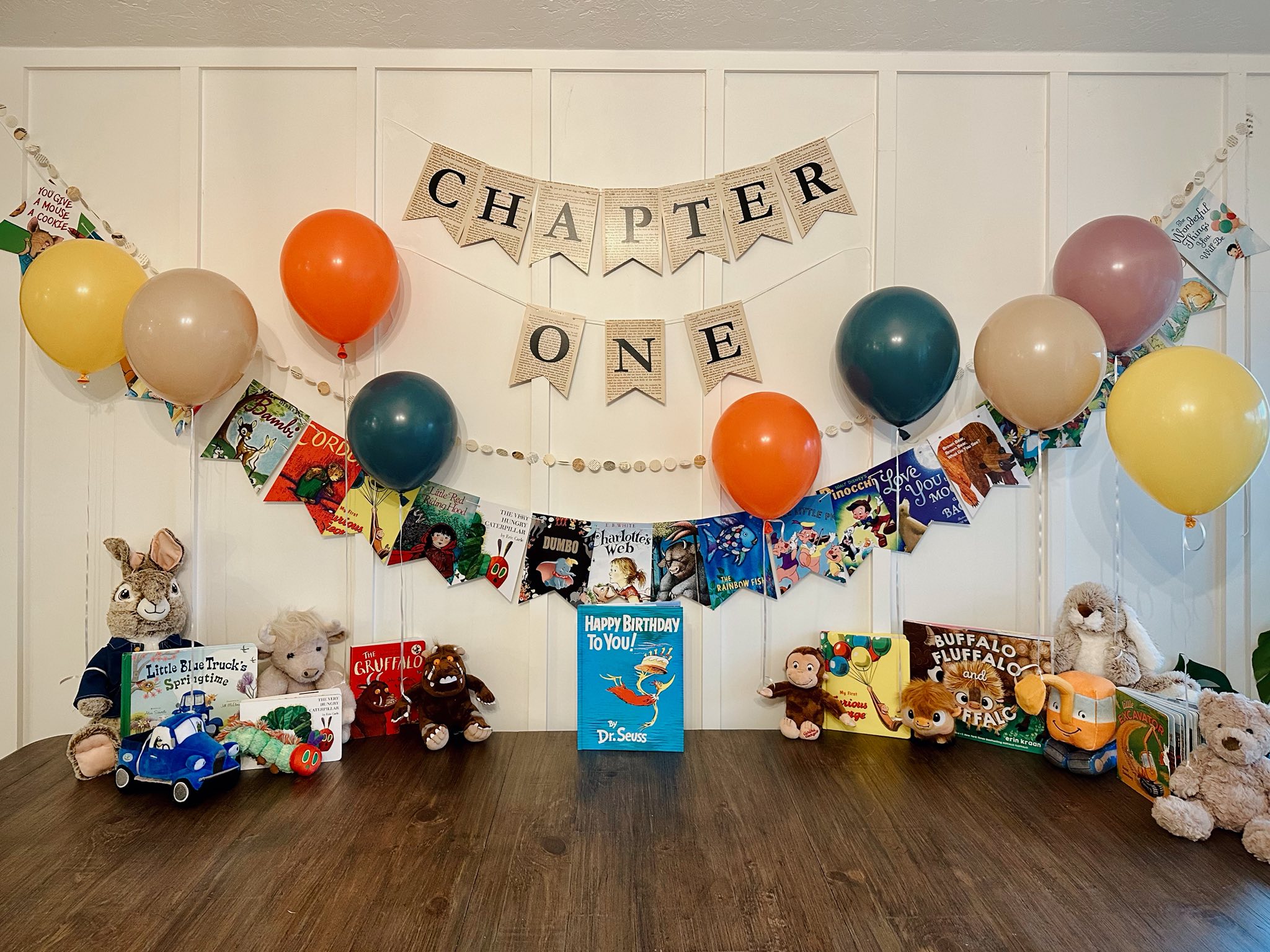 First birthday decorations featuring a Chapter One banner, mixed colored balloons, and a collection of classic children's books and plush toys.