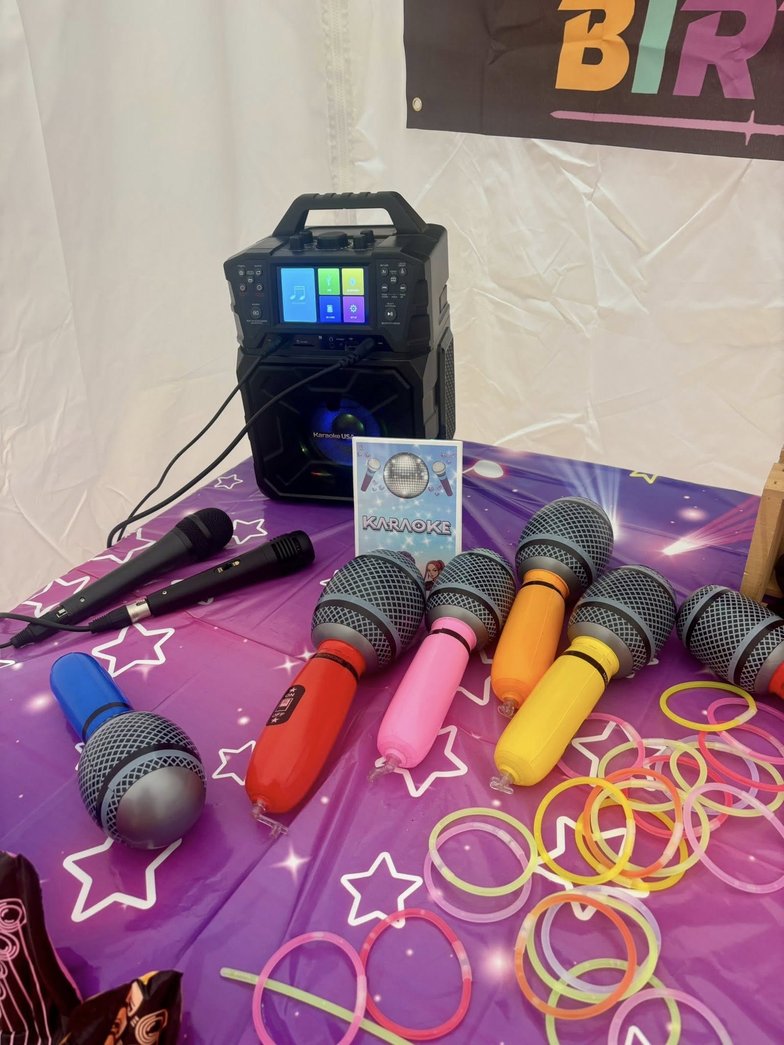 Karaoke machine with multiple colorful toy microphones and glow sticks spread on a purple star-patterned table.