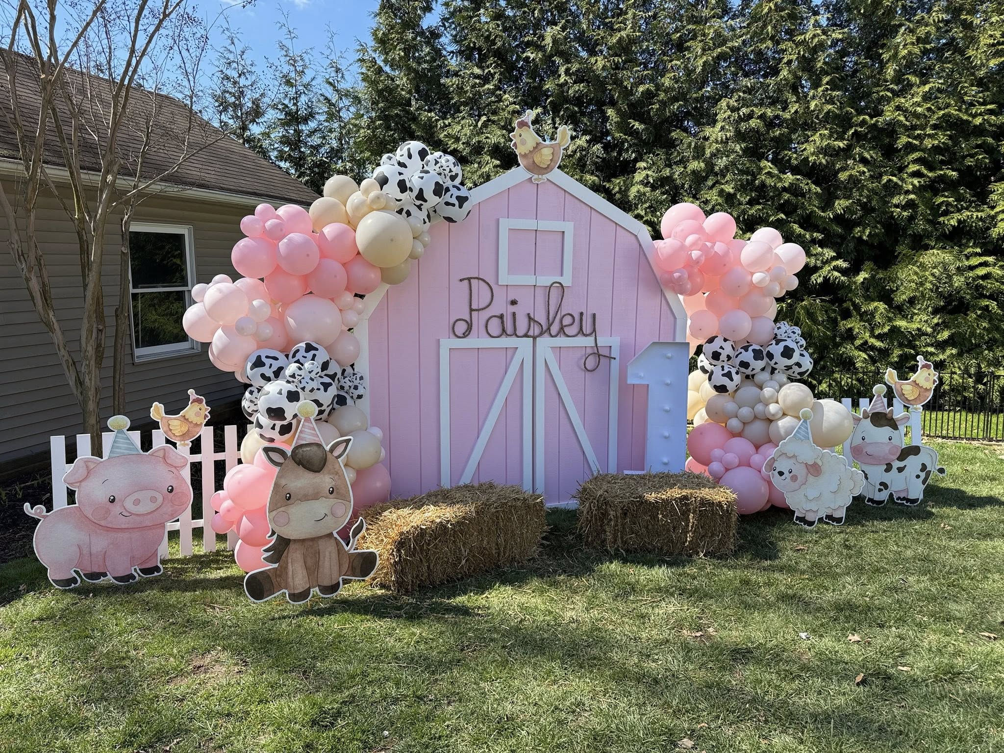 Pink barn backdrop for a first birthday party with cow print and pink balloon garland, hay bales, and farm animal cutouts.