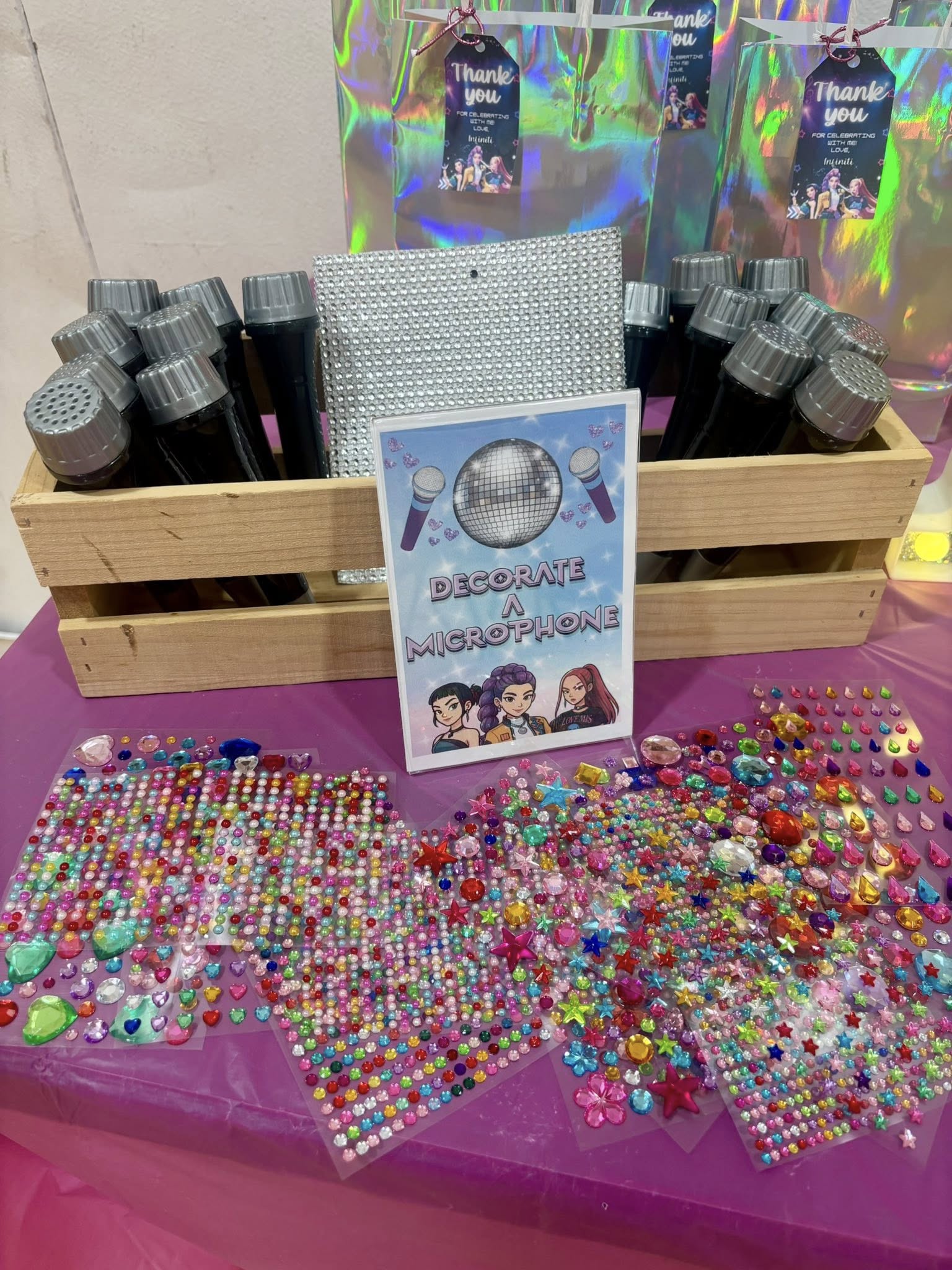 Table with a wooden crate of toy microphones and colorful adhesive rhinestone stickers at a DIY party station.