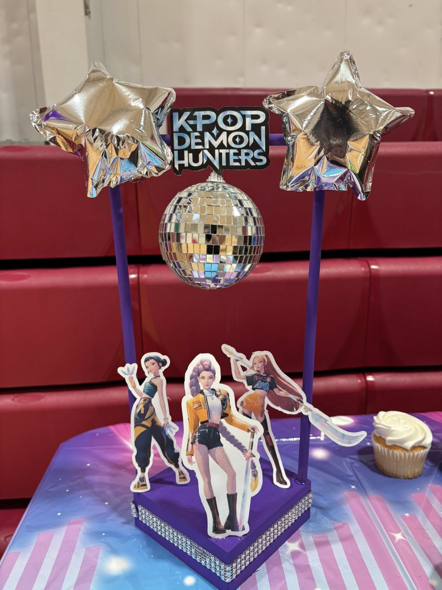 K-pop Demon Hunters party centerpiece with disco ball, star balloons, and cutouts of three characters.