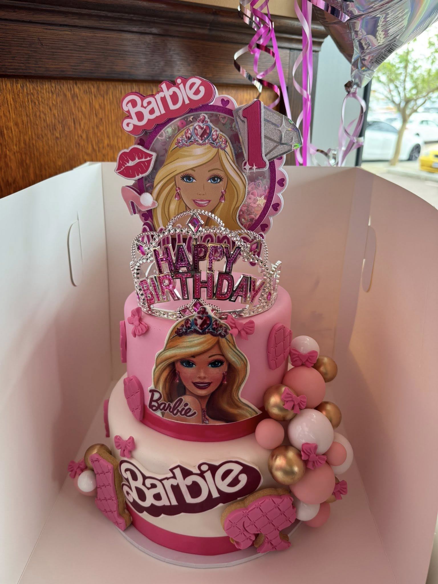 Two-tier pink Barbie-themed birthday cake with a crown topper, Barbie images, and pink and gold balloon decor.