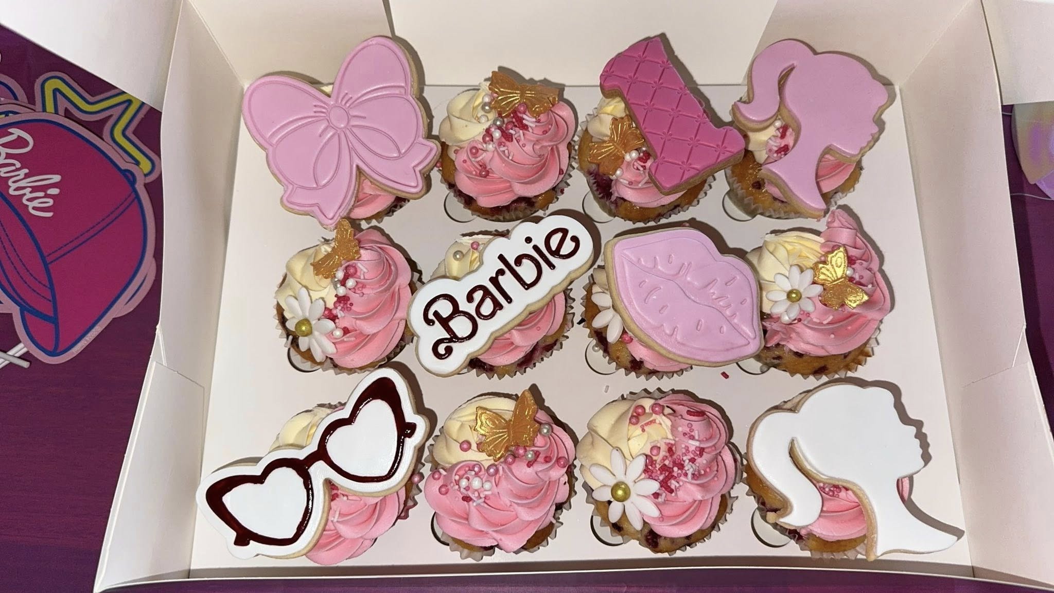 Box of twelve Barbie-themed cupcakes decorated with pink frosting, floral accents, and fondant cutouts.