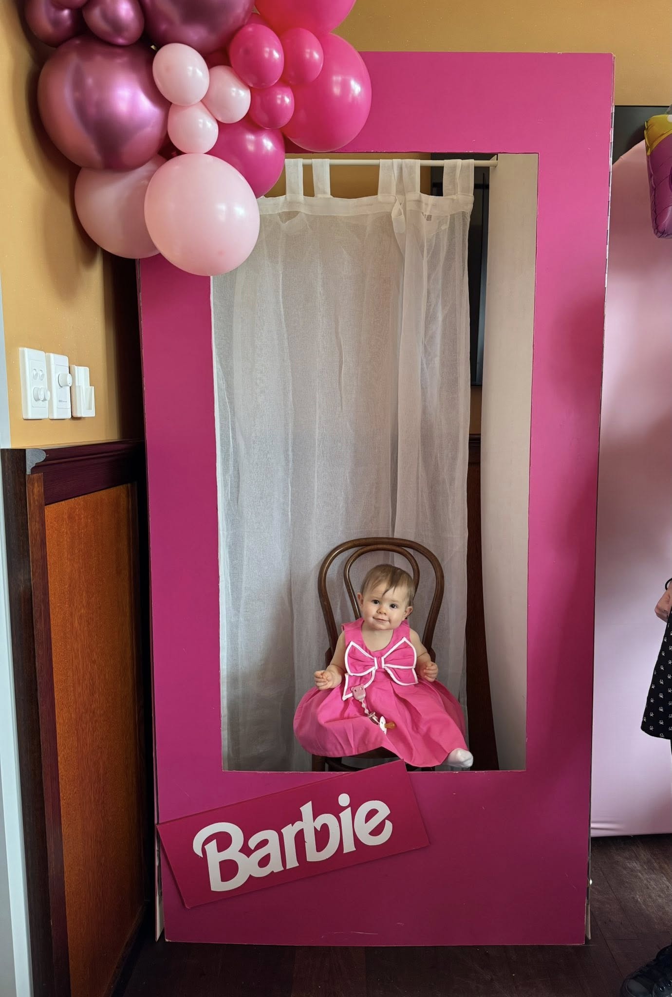 A baby girl in a pink dress sitting inside a giant, life-sized Barbie doll box photo booth prop.